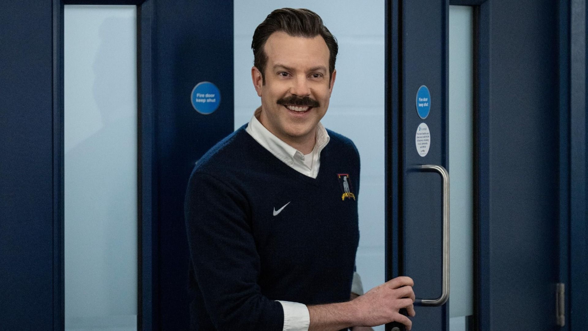 El actor Jason Sudeikis con un suéter azul y bigote parado cerca de una puerta sonriendo