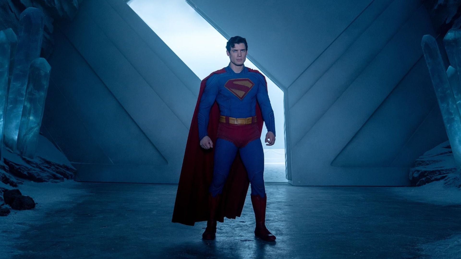 El actor David Corenswet con un traje de superhéroe azul y rojo junto a unas grandes puertas