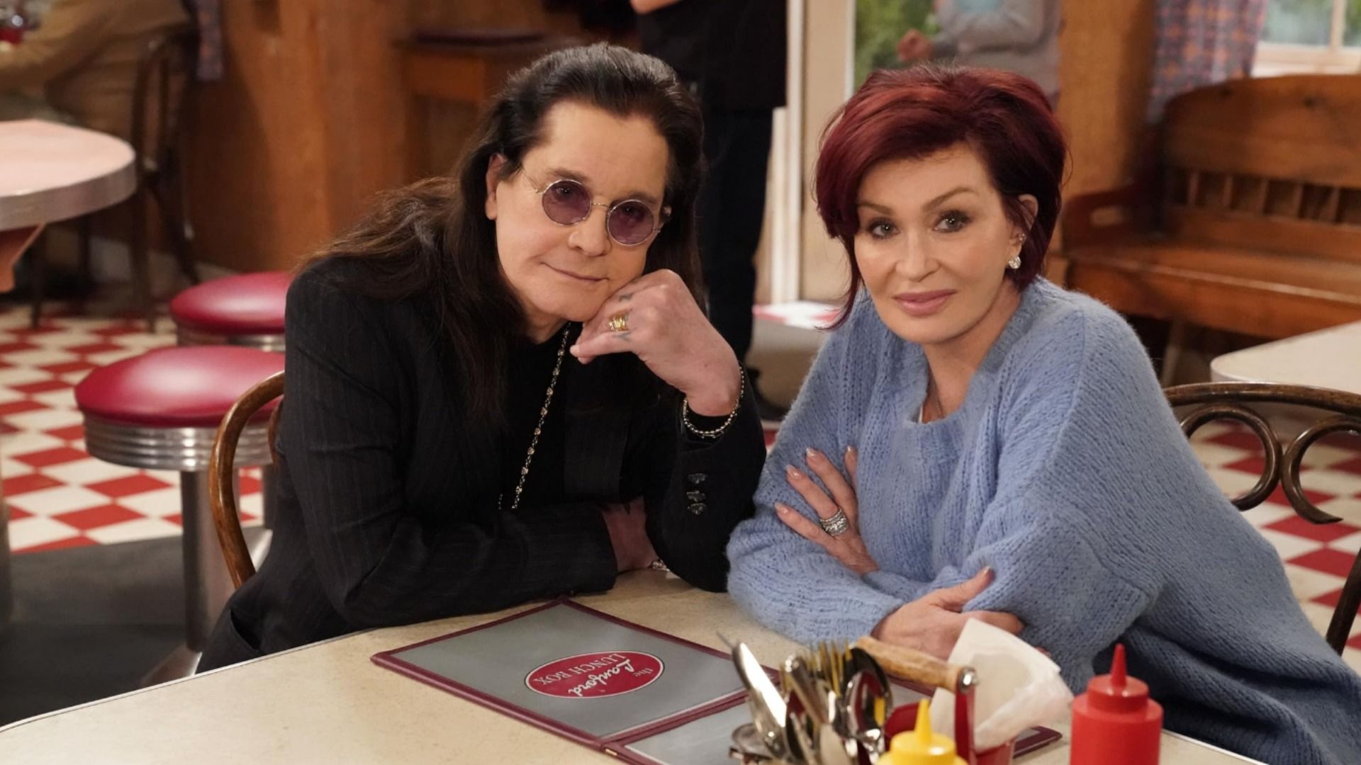 El cantante Ozzy Osbourne con gafas y chaqueta negra sentado en una mesa con su esposa