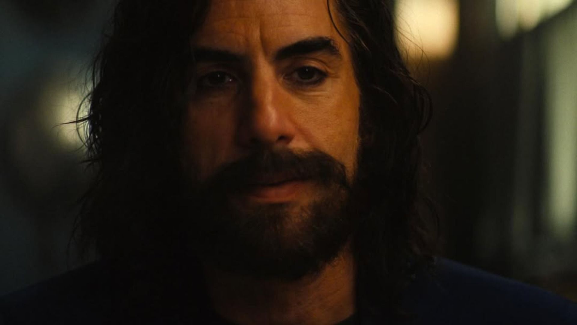 El actor Sacha Baron Cohen con barba y cabello largo está de pie contra un fondo borroso