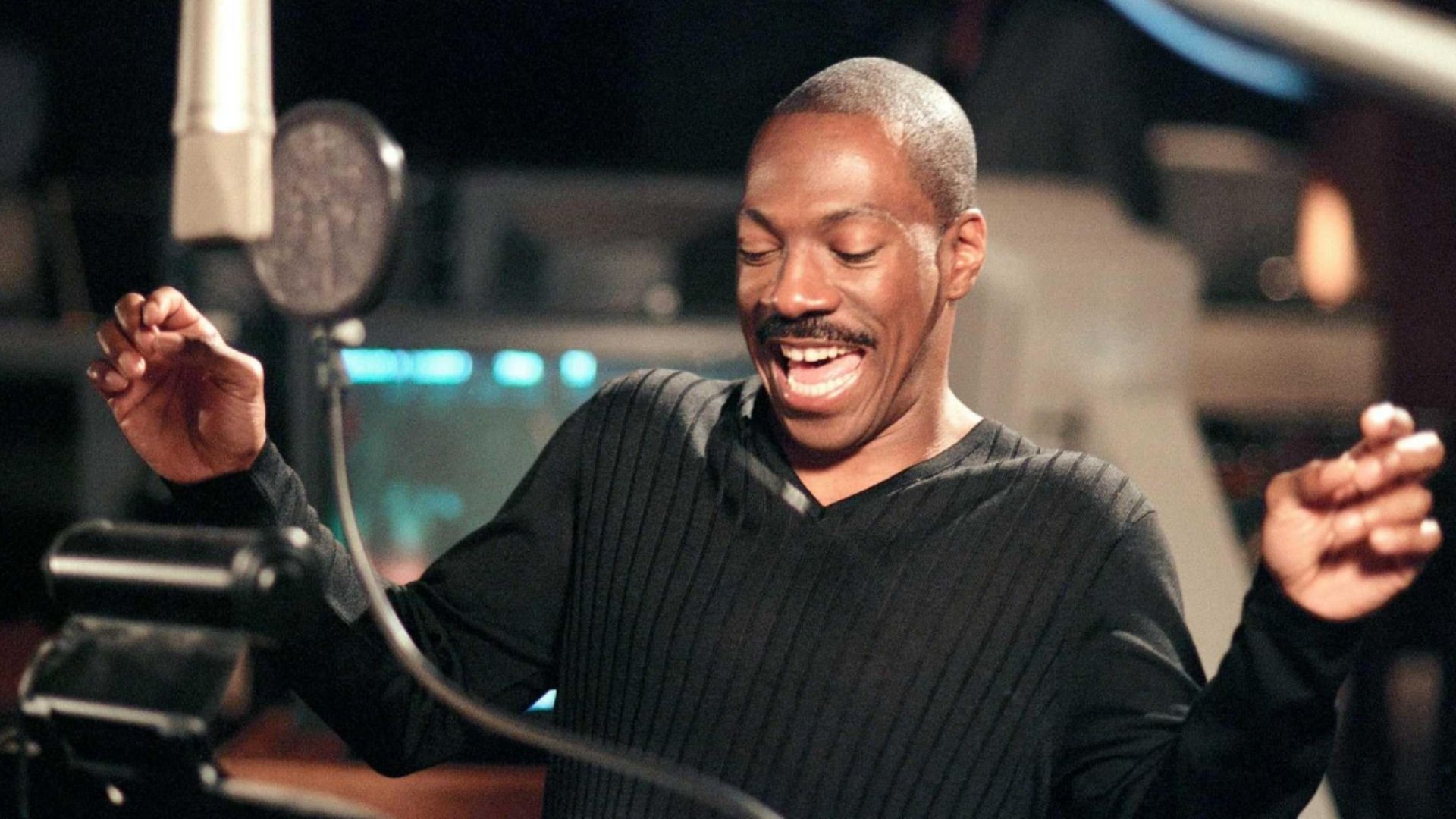 El actor Eddie Murphy con un suéter negro está parado frente a un micrófono y sonríe