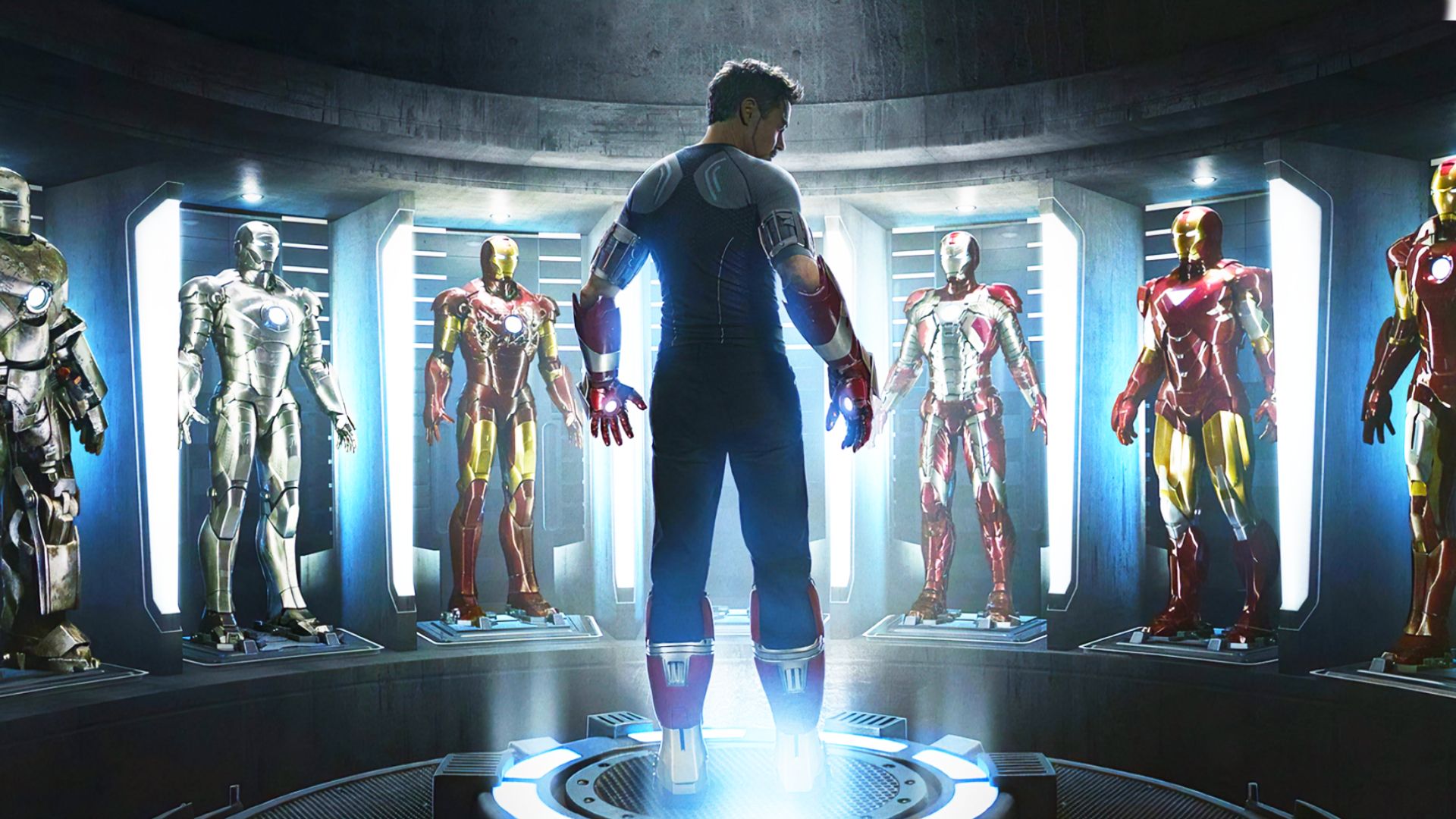 El actor Robert Downey Jr. vestido de gris y negro está rodeado de trajes de Iron Man