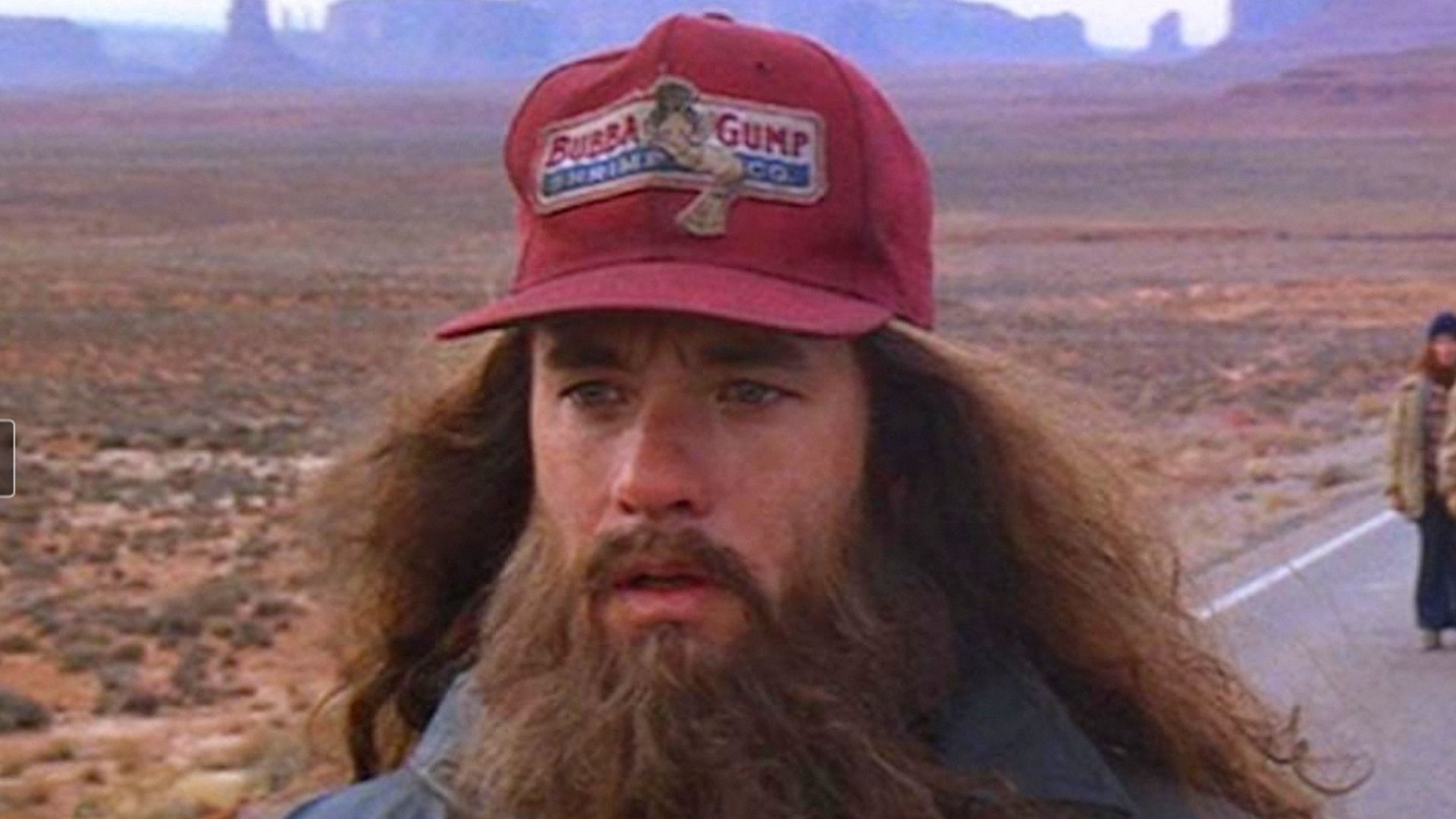 El actor Tom Hanks con cabello largo y barba de pie usando una gorra roja