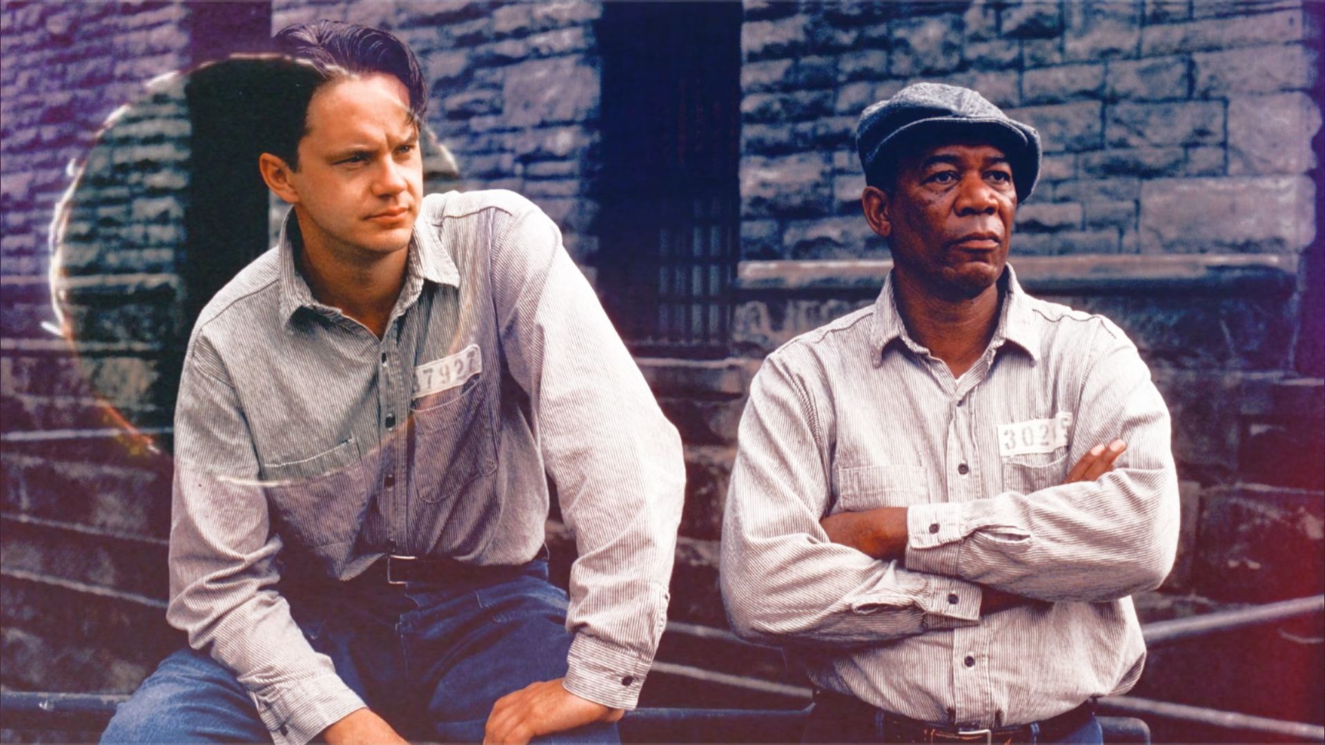 Los actores Tim Robbins y Morgan Freeman con uniformes de prisión sentados junto a un muro de piedra