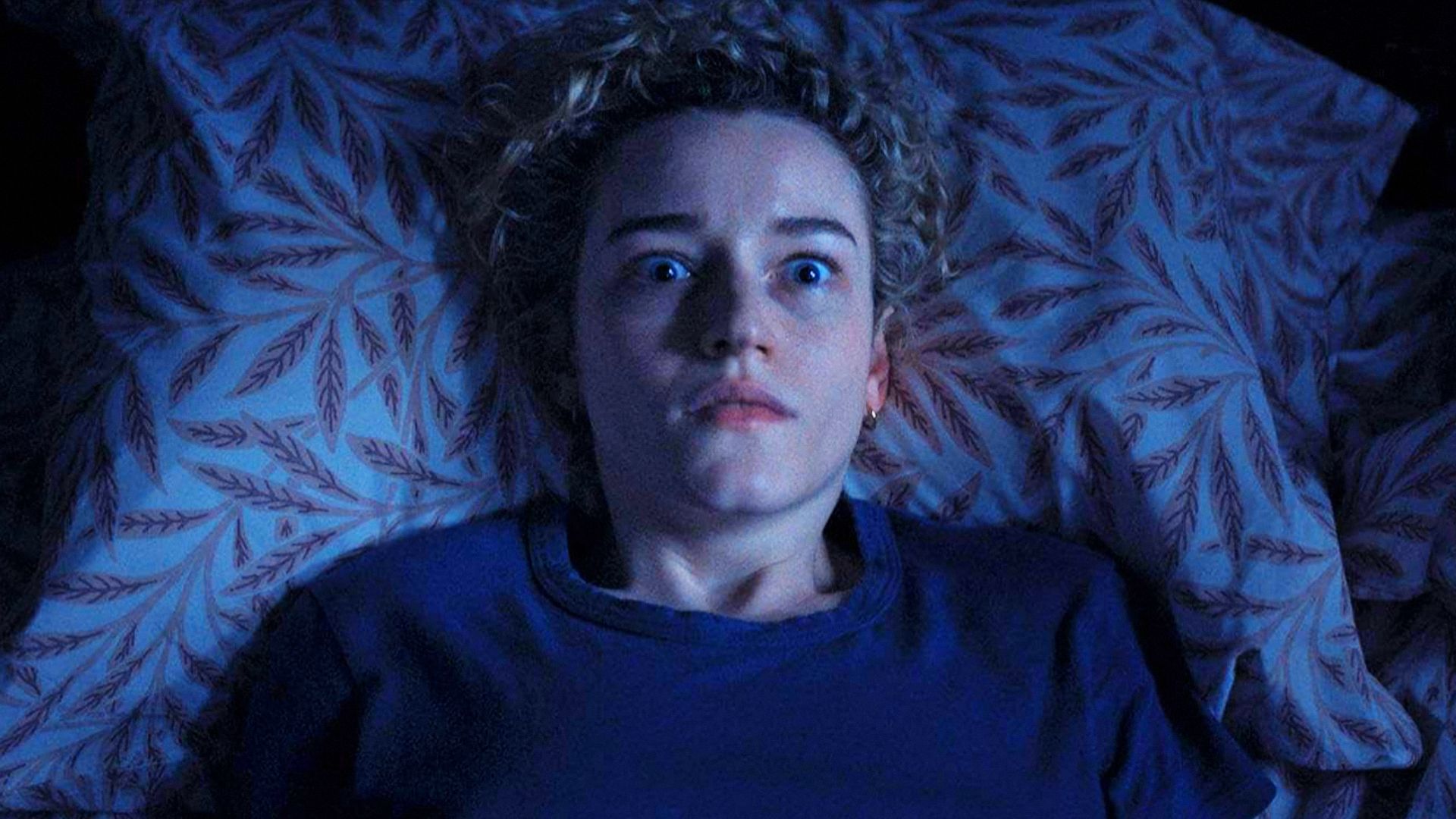 La actriz Julia Garner con un suéter azul yace en una cama, asustada por algo