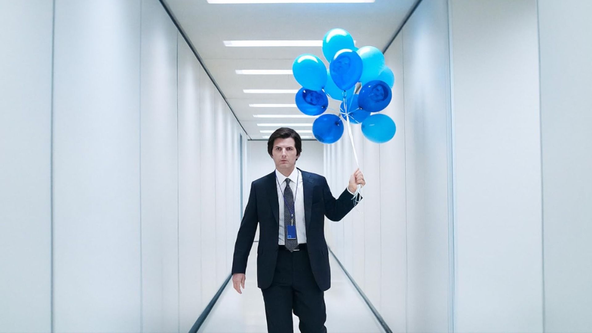 El actor Adam Scott con un traje clásico camina por un pasillo blanco sosteniendo globos azules