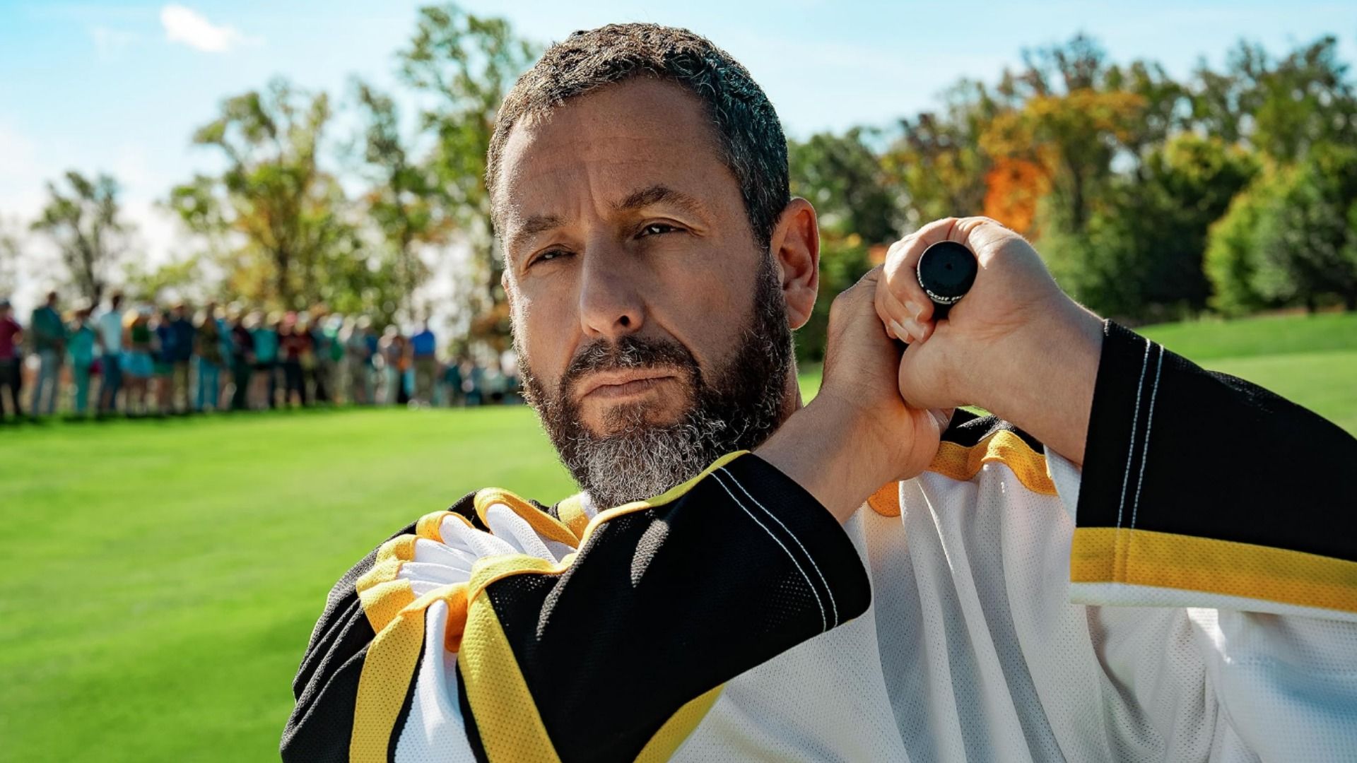 El actor Adam Sandler sosteniendo un palo de golf parado en un campo de golf