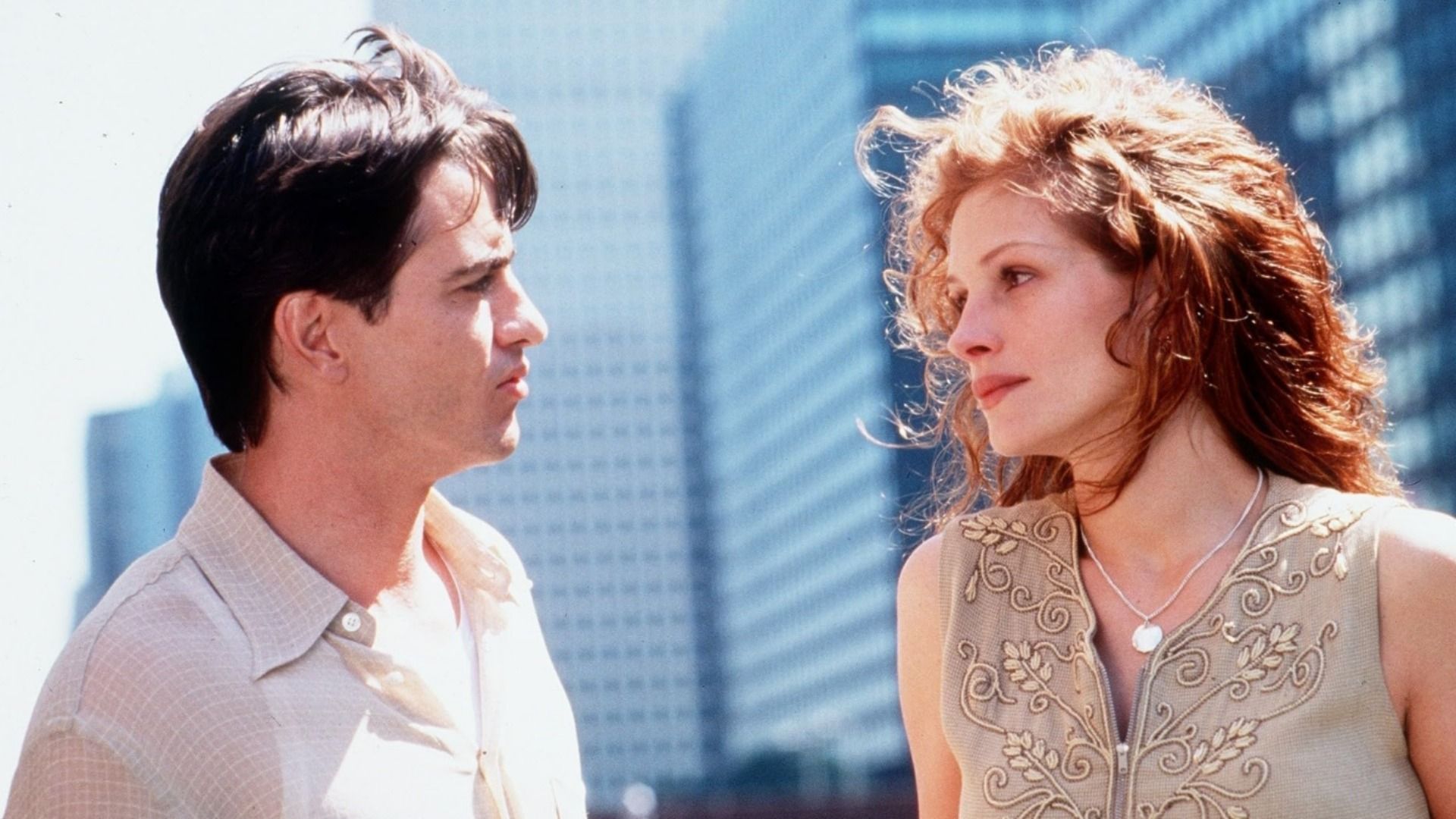 El actor Dermot Mulroney con camisa clara mira a la actriz Julia Roberts con rascacielos de fondo
