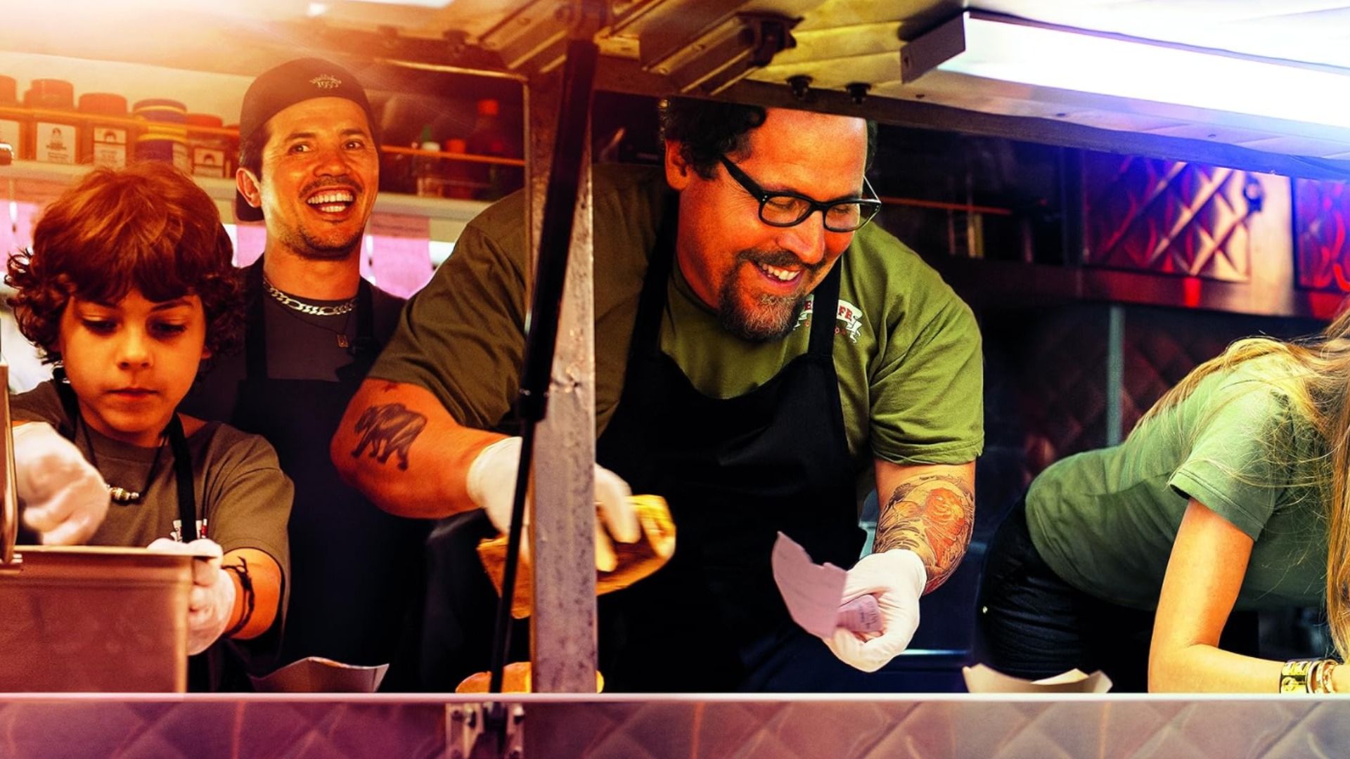 El actor Jon Favreau con delantal negro sostiene un panecillo rodeado de un niño, un hombre y una mujer