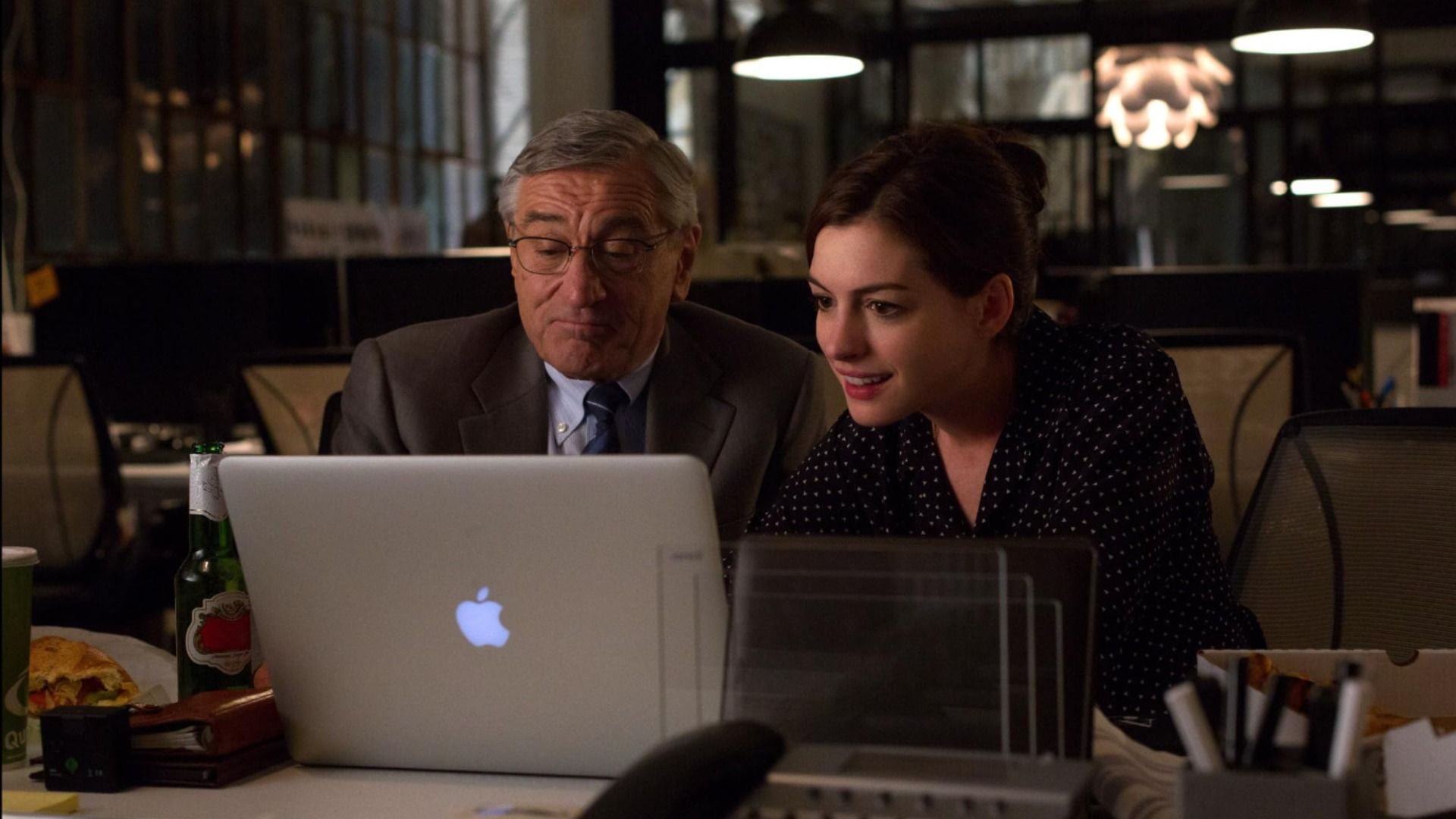 El actor Robert De Niro con chaqueta gris y la actriz Anne Hathaway con vestido negro sentados en una mesa