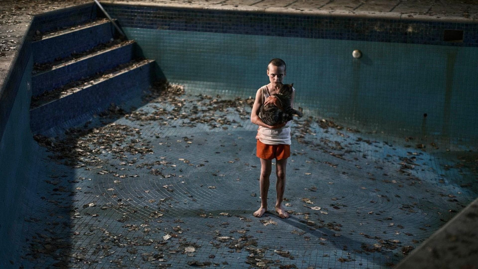 Un chico en pantalones naranjas está parado en el fondo de una piscina sosteniendo un gato