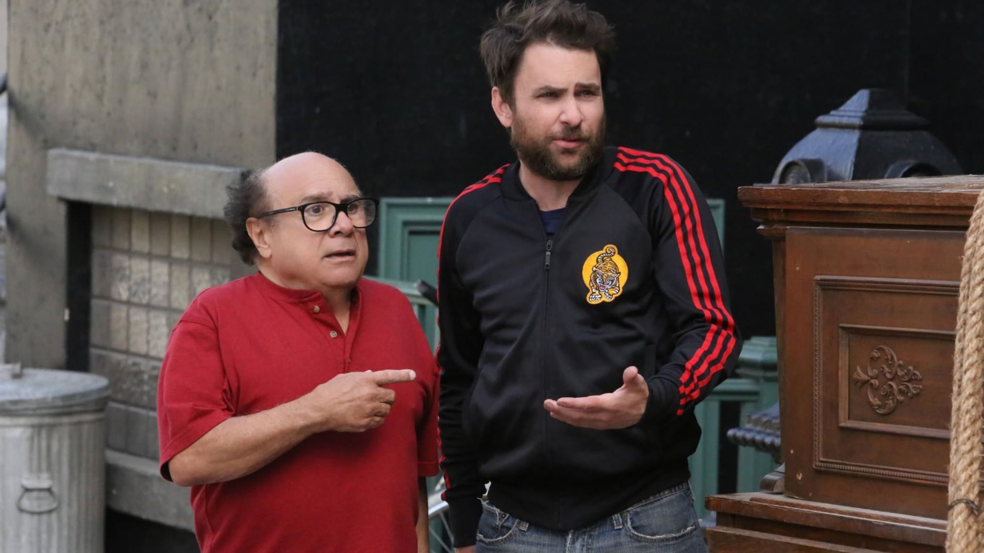 El actor Danny DeVito en camisa roja señala con el dedo a un hombre en ropa deportiva