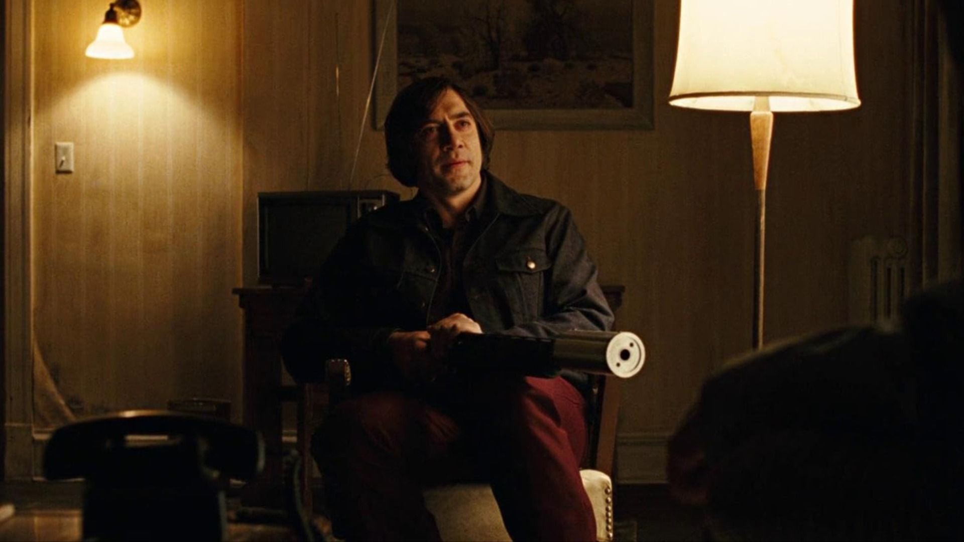 El actor Javier Bardem con pelo largo sentado en una silla sosteniendo un arma