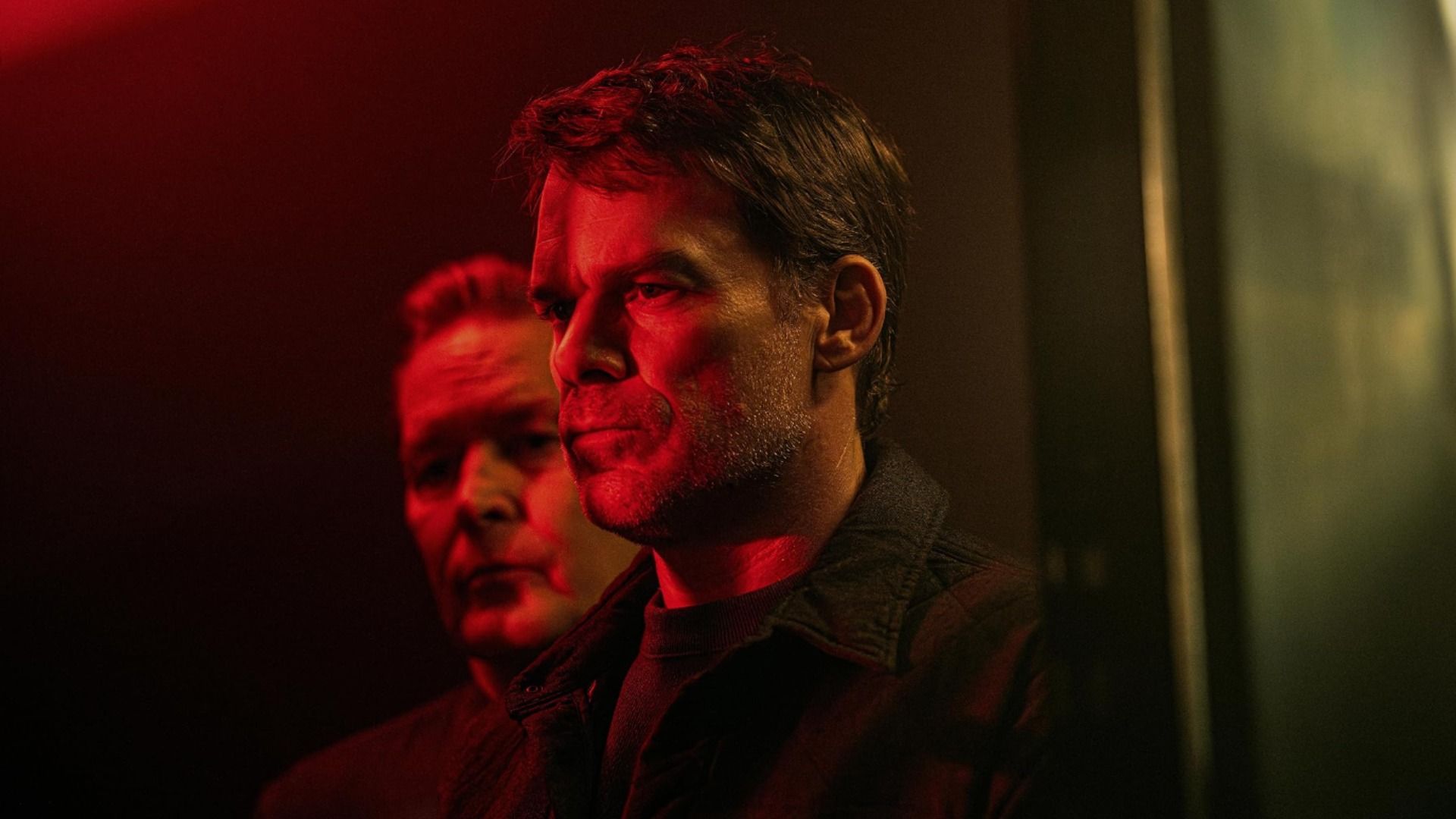 El actor Michael C. Hall se encuentra de perfil bajo iluminación roja con parte del rostro de un hombre visible detrás de él