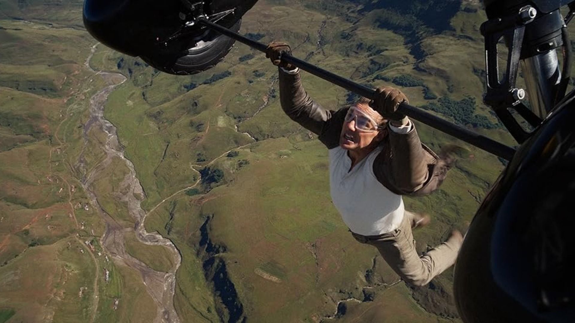 El actor Tom Cruise con chaqueta marrón se agarra a las ruedas de un avión en vuelo