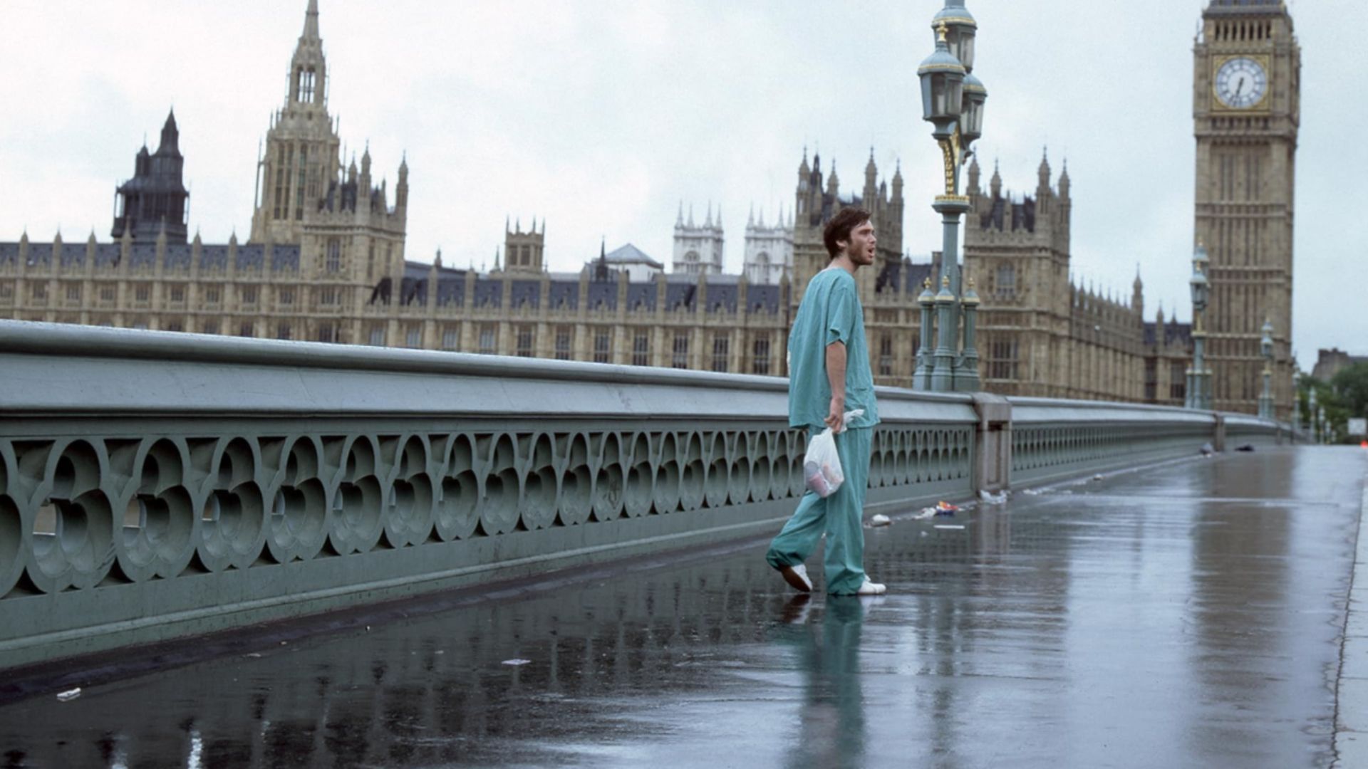 El actor Cillian Murphy con ropa azul está de pie en un puente vacío con la torre del reloj Big Ben de Londres visible cerca