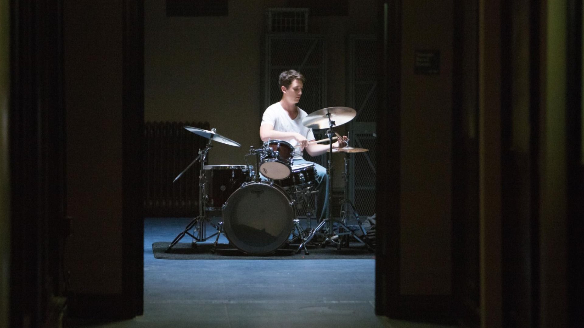 Iluminación tenue, el actor Miles Teller toca la batería con una camiseta blanca