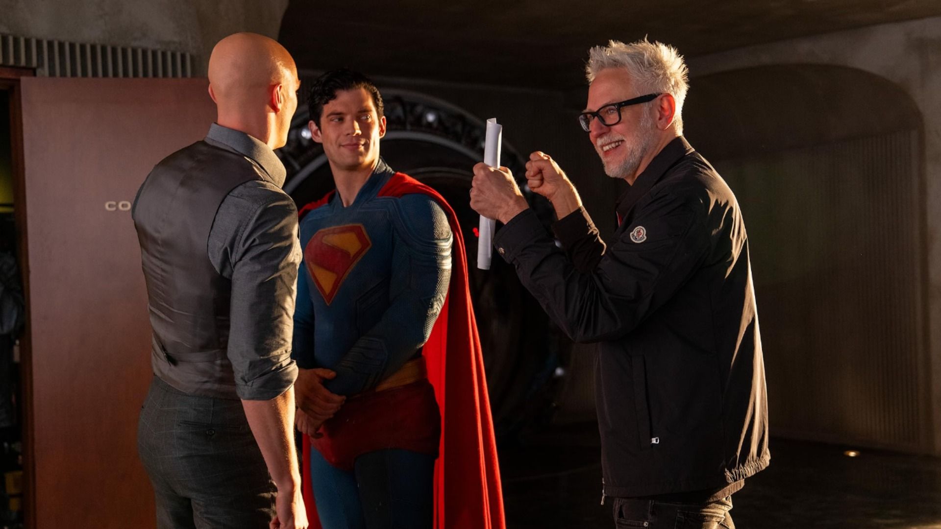 Director James Gunn sosteniendo papeles habla con el actor David Corenswet en traje de Superman y con Nicholas Hoult que está calvo