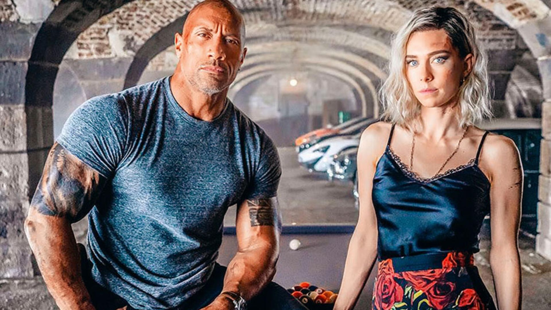 Dwayne Johnson con una camiseta ajustada y Vanessa Kirby con una camiseta sin mangas y falda estampada posan contra un fondo de arcos y coches deportivos