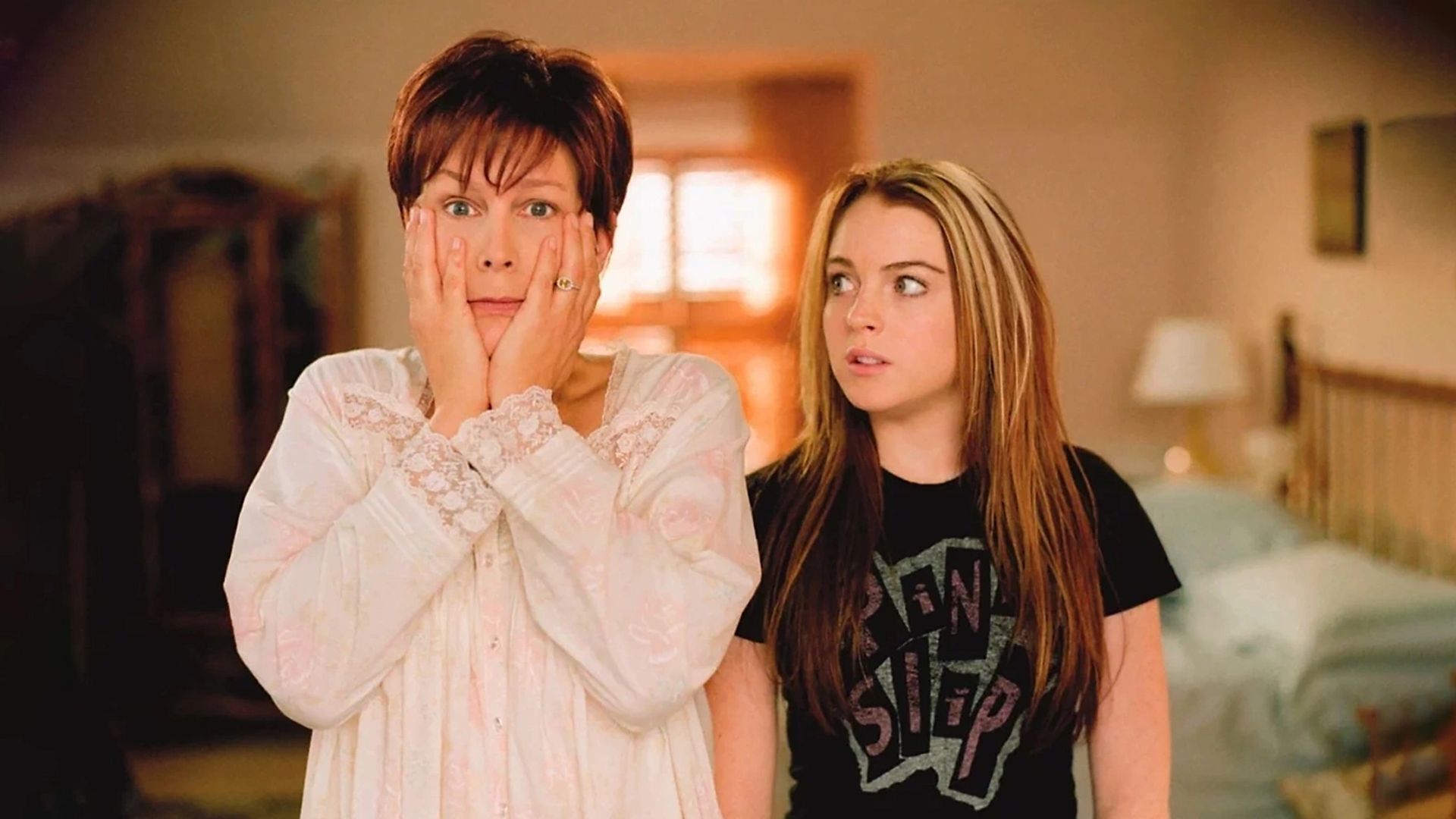 Jamie Lee Curtis en pijama sorprendida con las manos cubriéndose la cara mientras Lindsay Lohan la mira con sorpresa