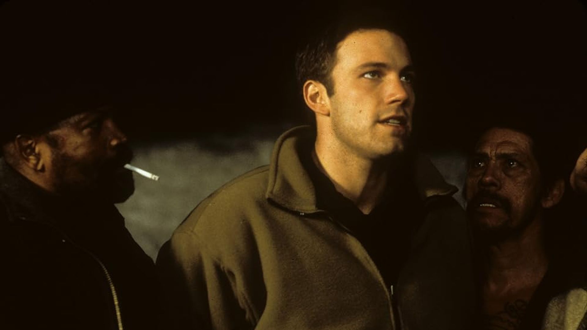 Ben Affleck con una chaqueta beige parado entre dos personas, una de las cuales está fumando, en un ambiente poco iluminado