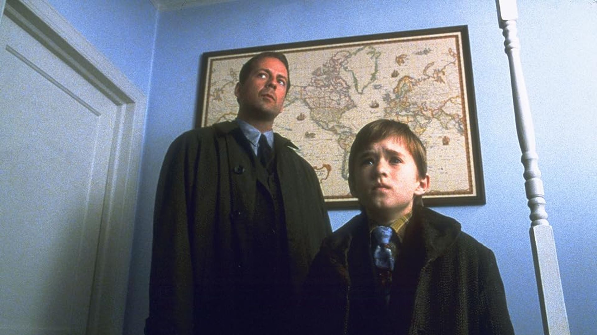 Bruce Willis y Haley Joel Osment de pie en un pasillo con un mapamundi colgado en la pared detrás de ellos