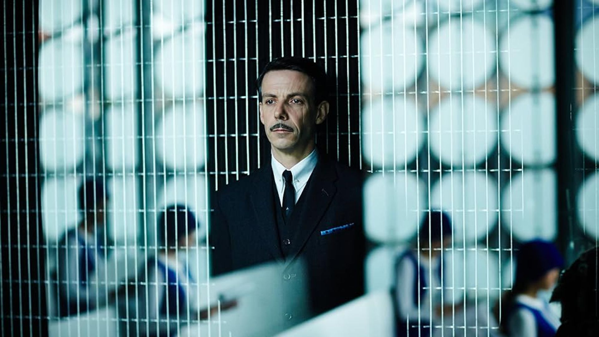 Ethan Hawke con traje y bigote parado detrás de una división de malla con gente uniformada trabajando en escritorios al fondo