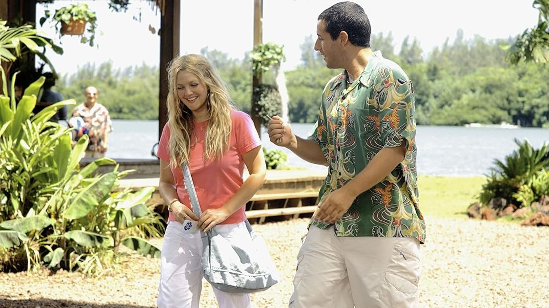 Drew Barrymore y Adam Sandler conversando en una playa soleada con vegetación y agua de fondo