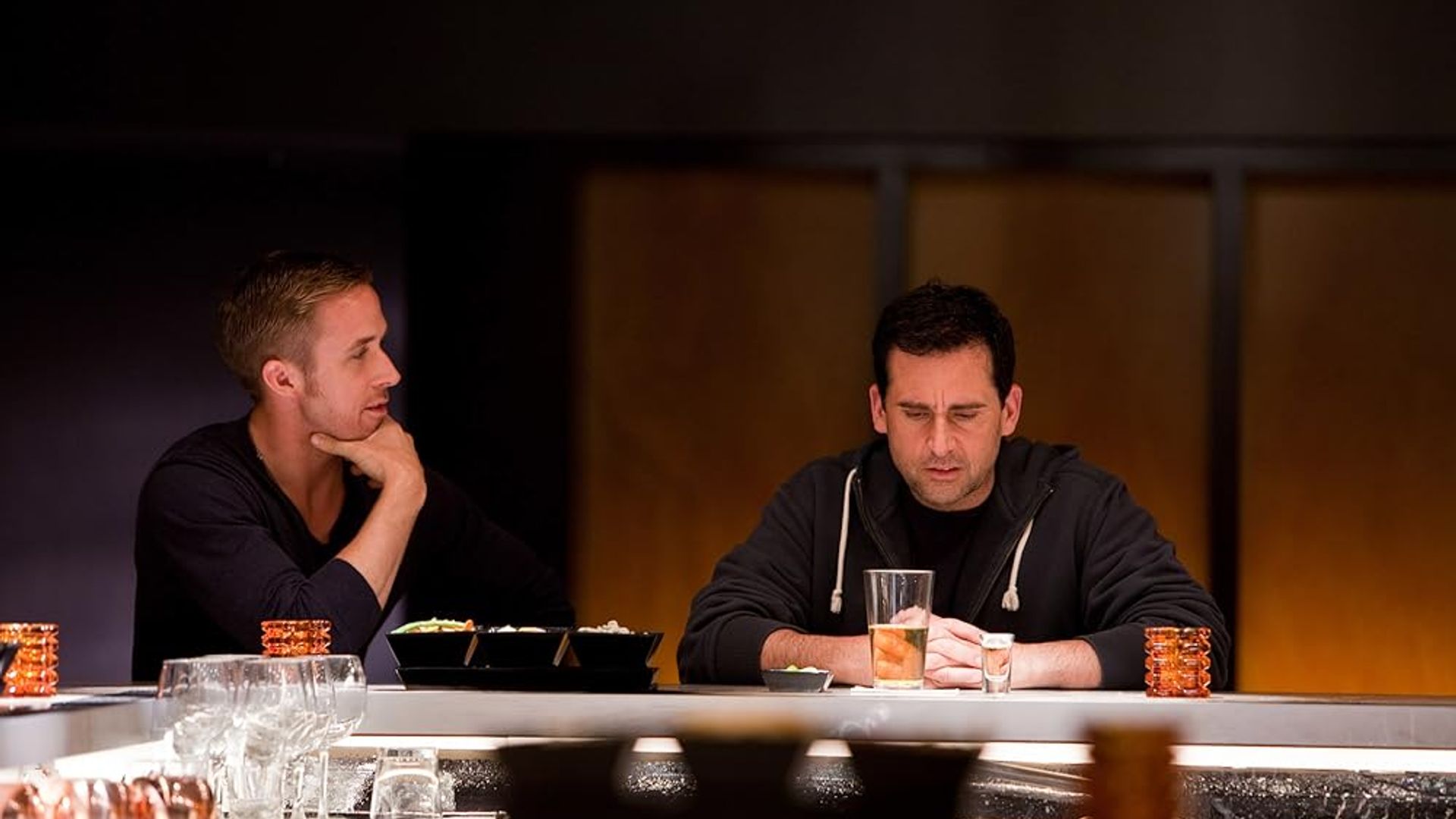 Ryan Gosling y Steve Carell sentados en la barra de un bar, uno mirando pensativo su copa, el otro observando