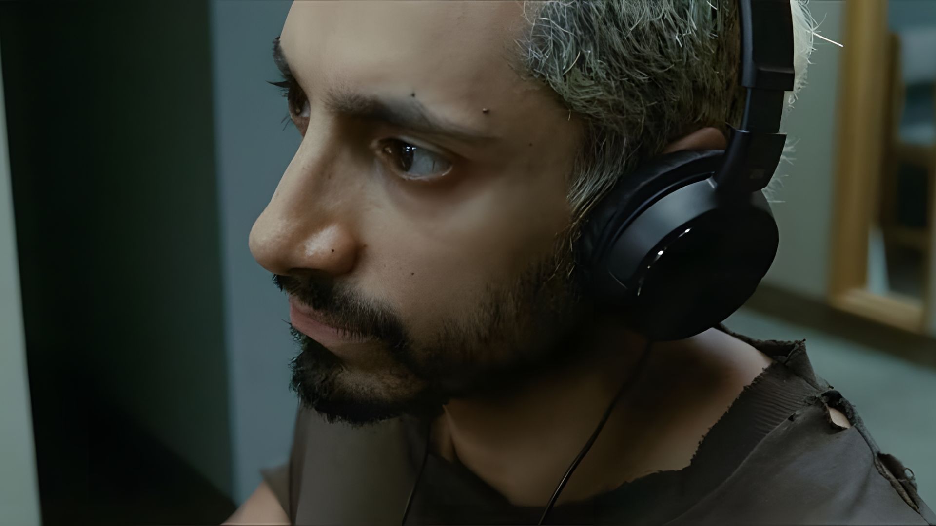 Riz Ahmed con auriculares y expresión seria sentado en interiores, vistiendo una camiseta rota