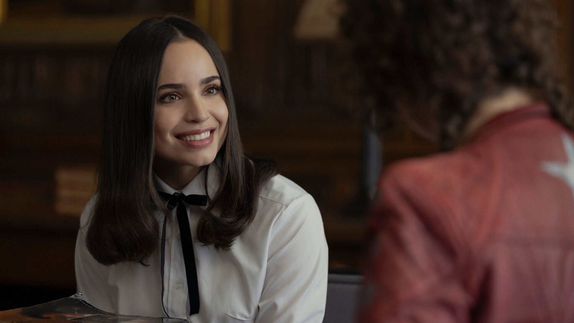 Sofia Carson con una blusa blanca y un lazo negro en el cuello sonriendo ampliamente