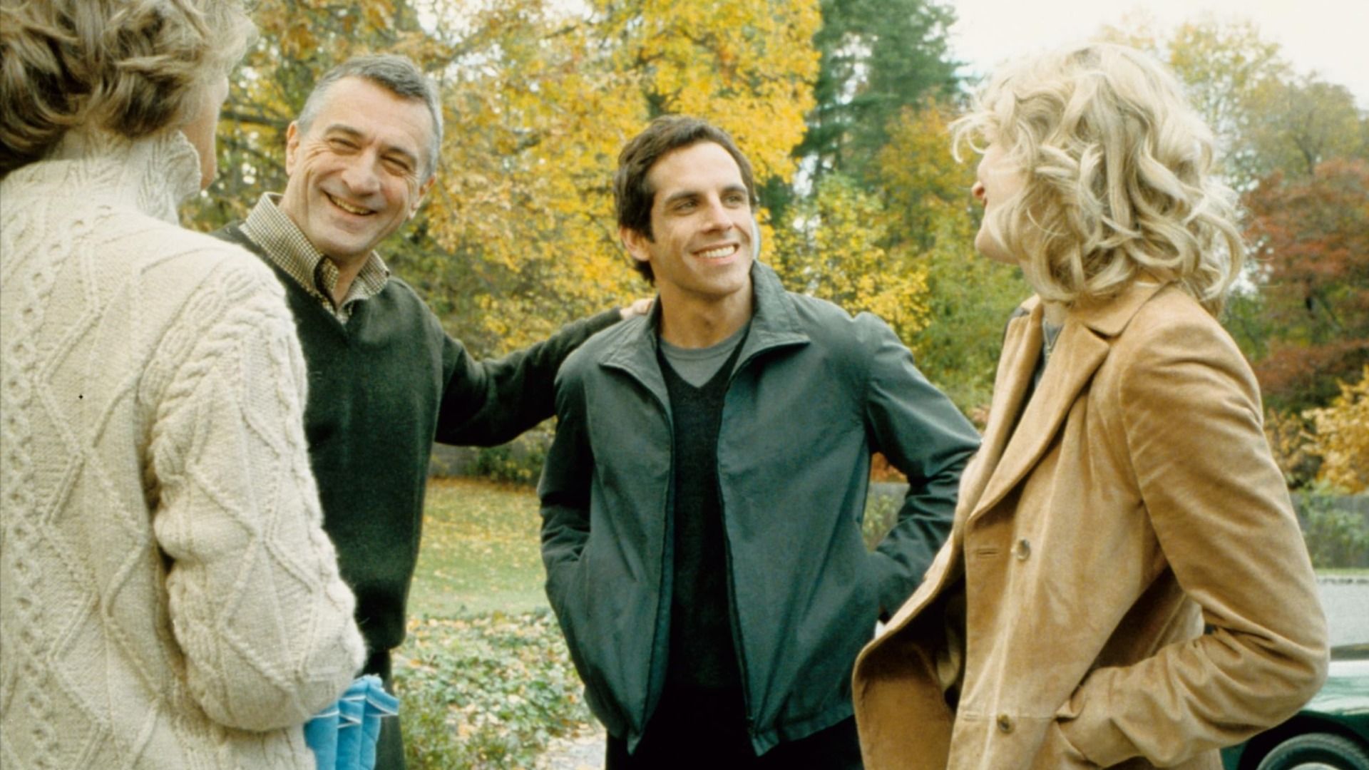 Los actores Robert De Niro con suéter verde y Ben Stiller con chaqueta verde junto a árboles con hojas amarillas