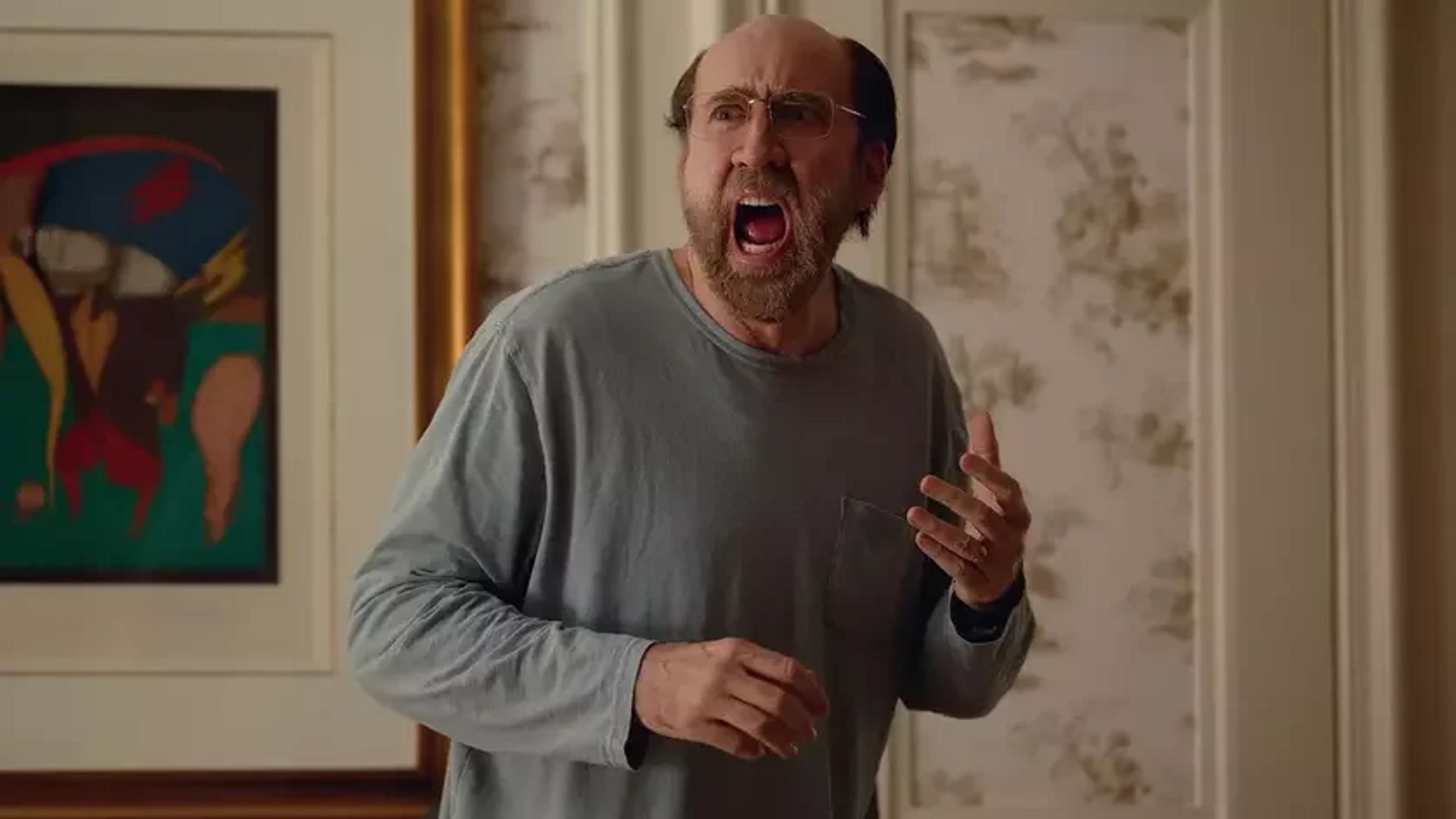Nicolas Cage con gafas y camisa gris de manga larga gritando mientras está de pie en una habitación