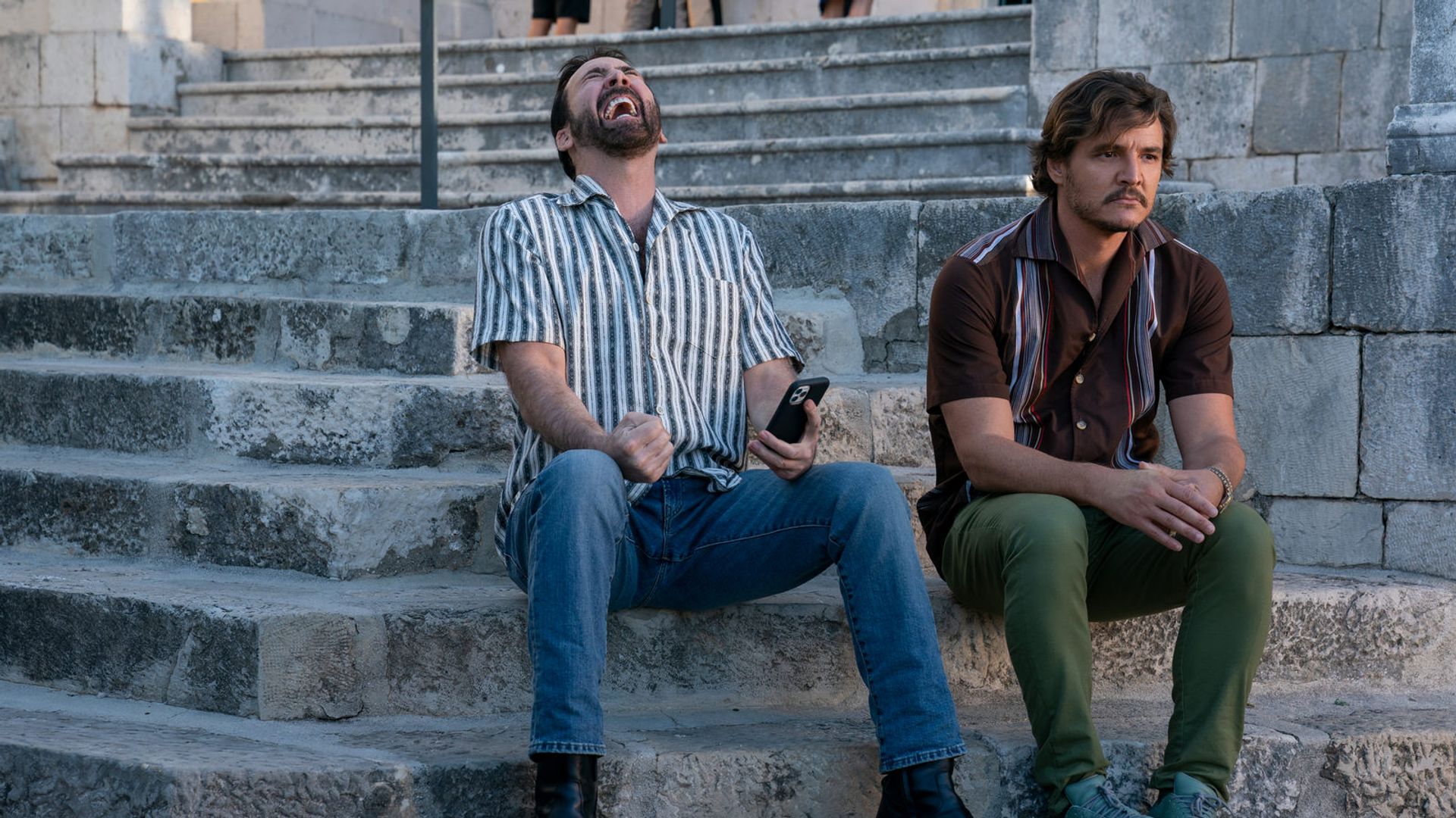 Nicolas Cage y Pedro Pascal sentados en escalones de piedra: uno ríe con un teléfono en la mano, el otro mira pensativamente hacia un lado