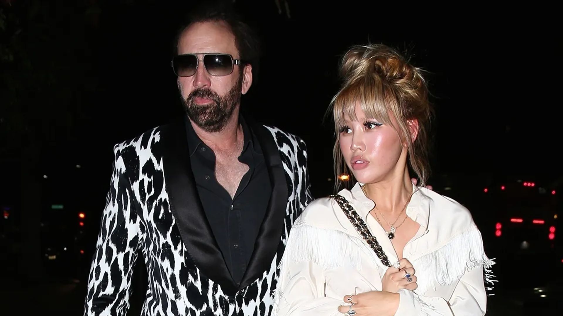 Nicolas Cage con un blazer estampado en blanco y negro y Erika Koike con una blusa blanca caminando juntos por la noche