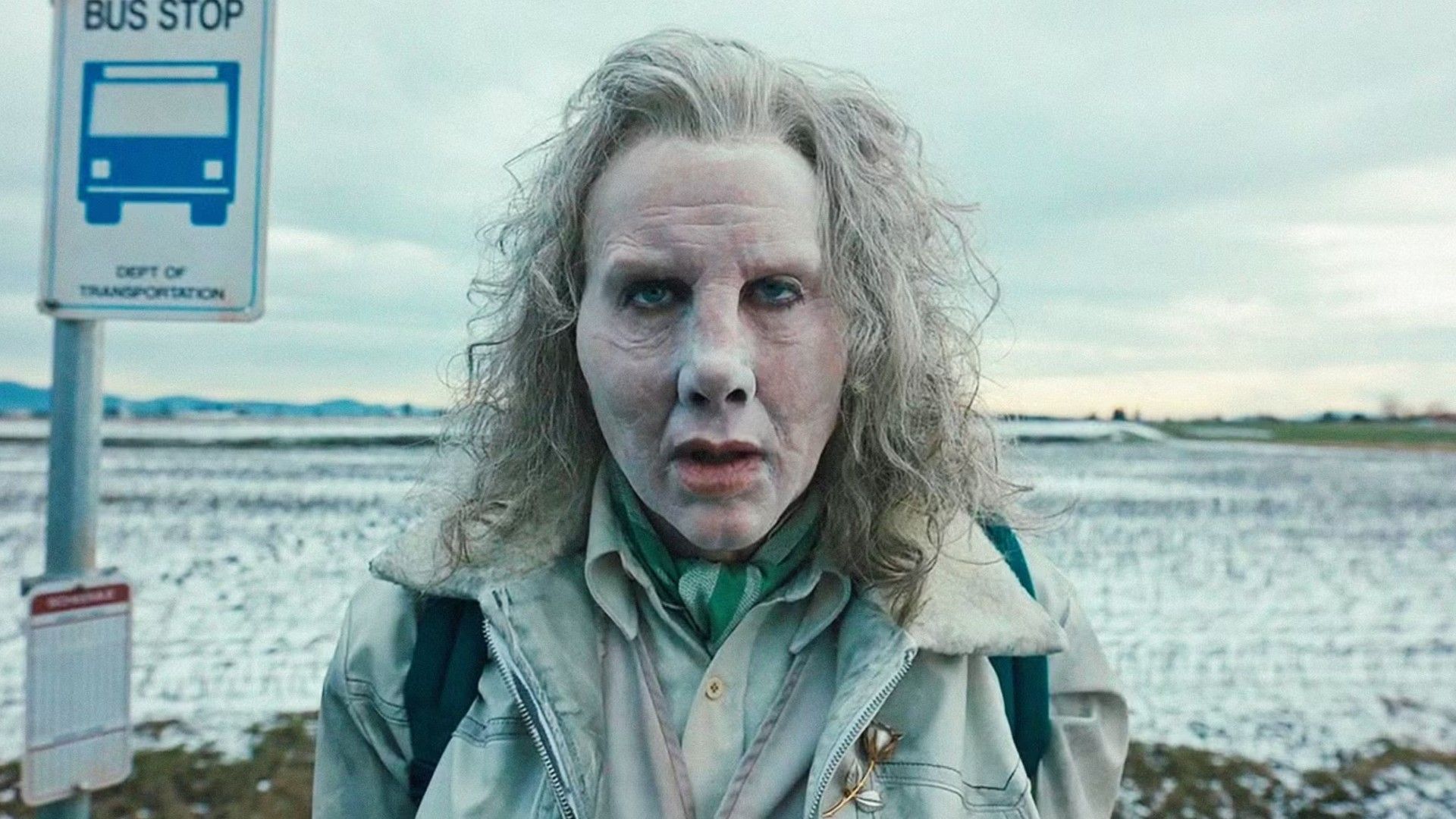 Nicolas Cage con cabello gris y rostro pálido junto a una señal de parada de autobús contra un paisaje invernal de fondo