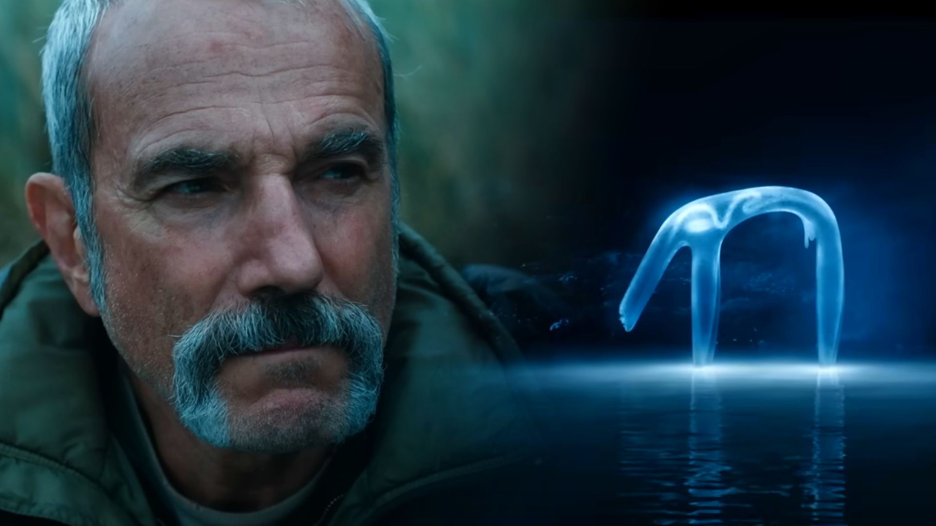 El actor Daniel Day-Lewis con chaqueta verde y una criatura azulada translúcida parada sobre el agua