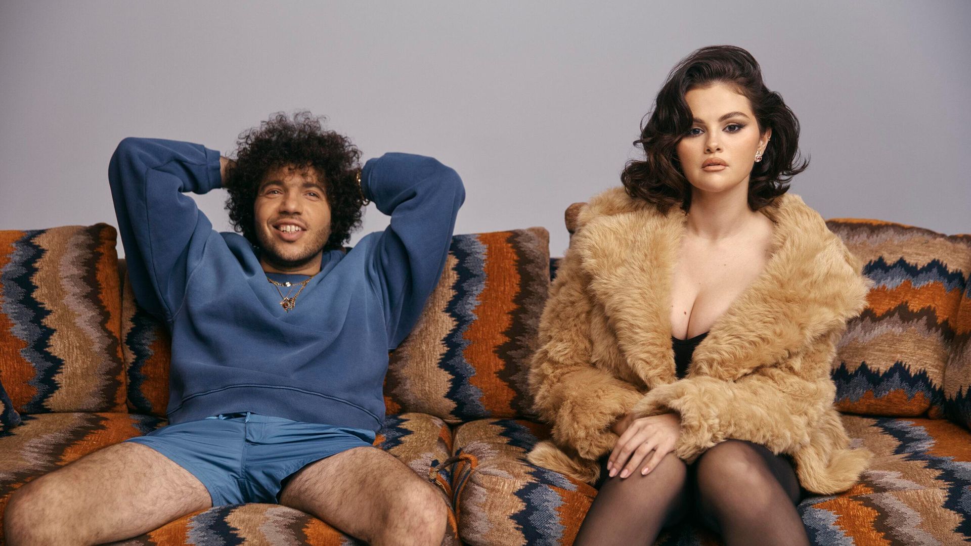 Selena Gomez con abrigo de piel y Benny Blanco con sudadera azul y shorts sentados relajados