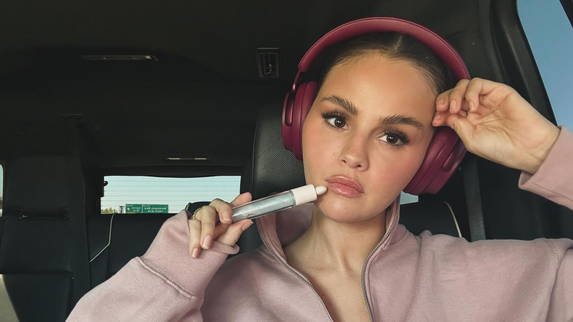 Selena Gomez con una sudadera rosa y auriculares sentada en un auto, sosteniendo un producto cosmético cerca de su cara