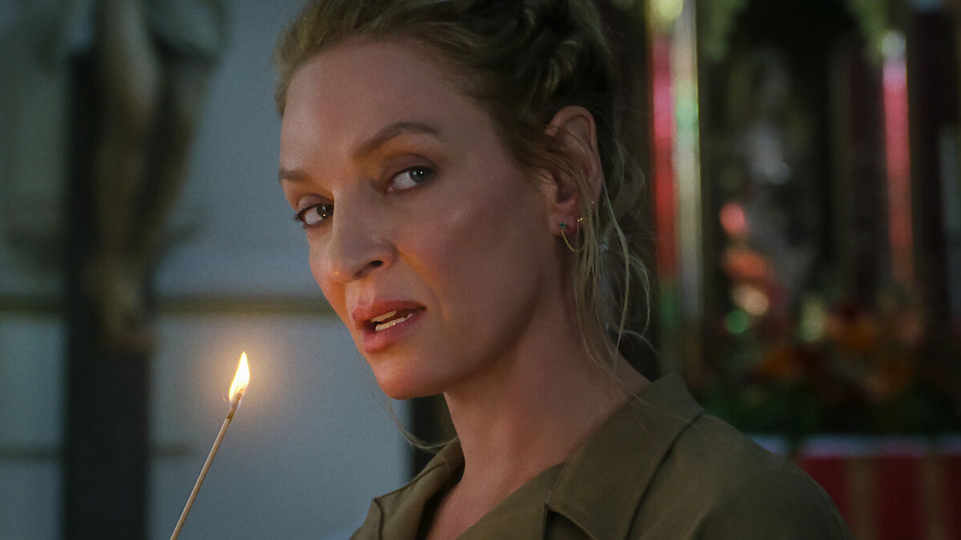 Uma Thurman con el cabello recogido sostiene una vela encendida y mira hacia un lado con un fondo ligeramente difuminado