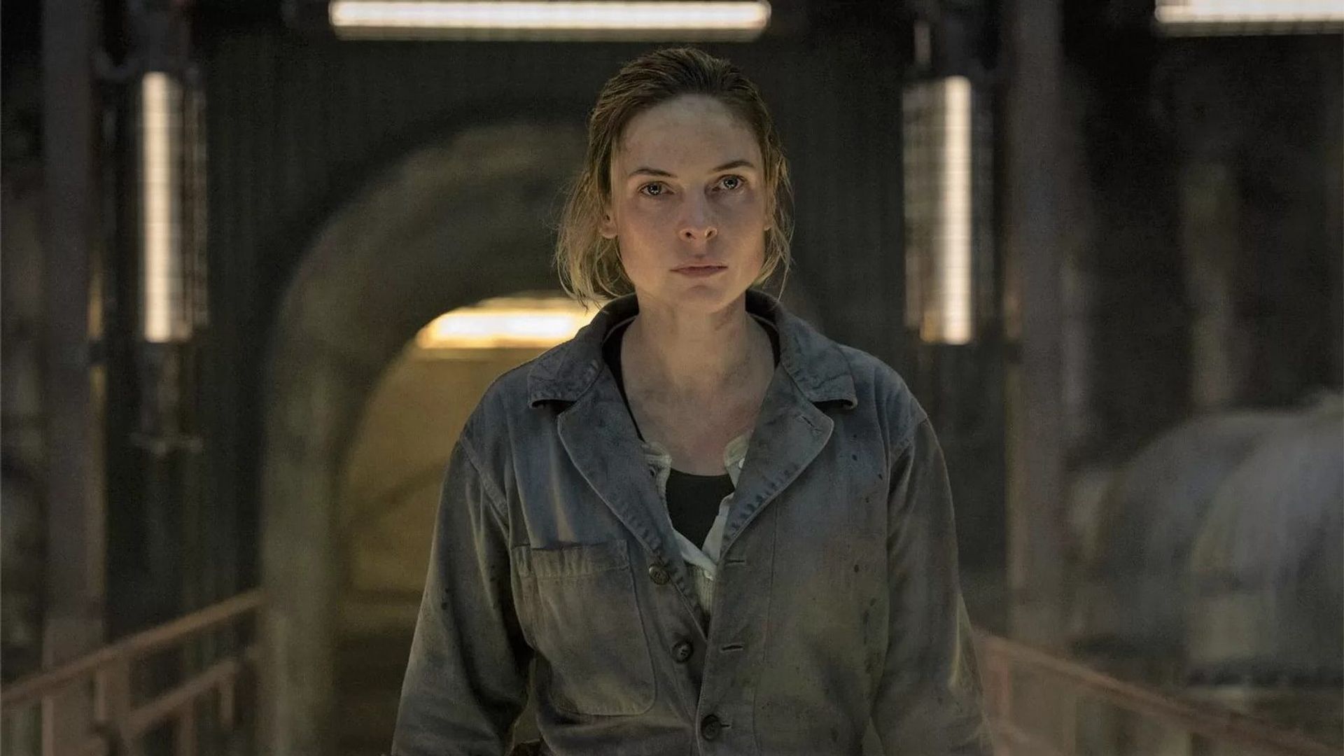 Rebecca Ferguson en overol de trabajo parada en un espacio industrial tenuemente iluminado, mirando directamente al frente