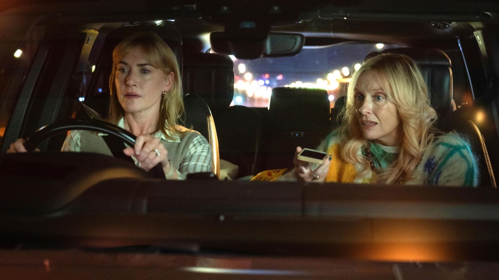 La actriz Kate Winslet sentada al volante de un coche mientras la actriz Toni Collette habla por teléfono a su lado
