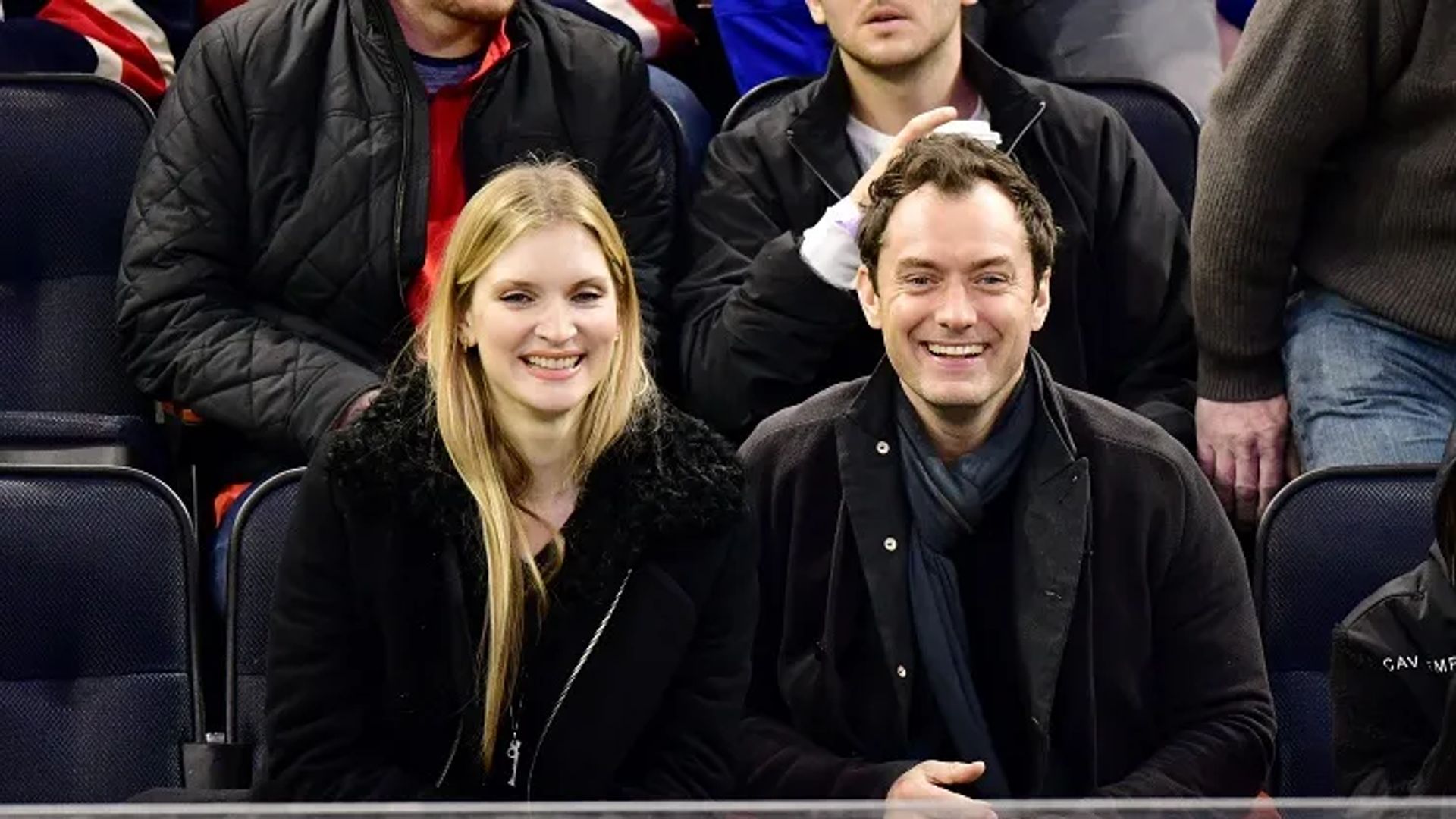 Jude Law y Phillipa Coan sentados uno al lado del otro en las gradas, sonriendo y mirando a la cámara