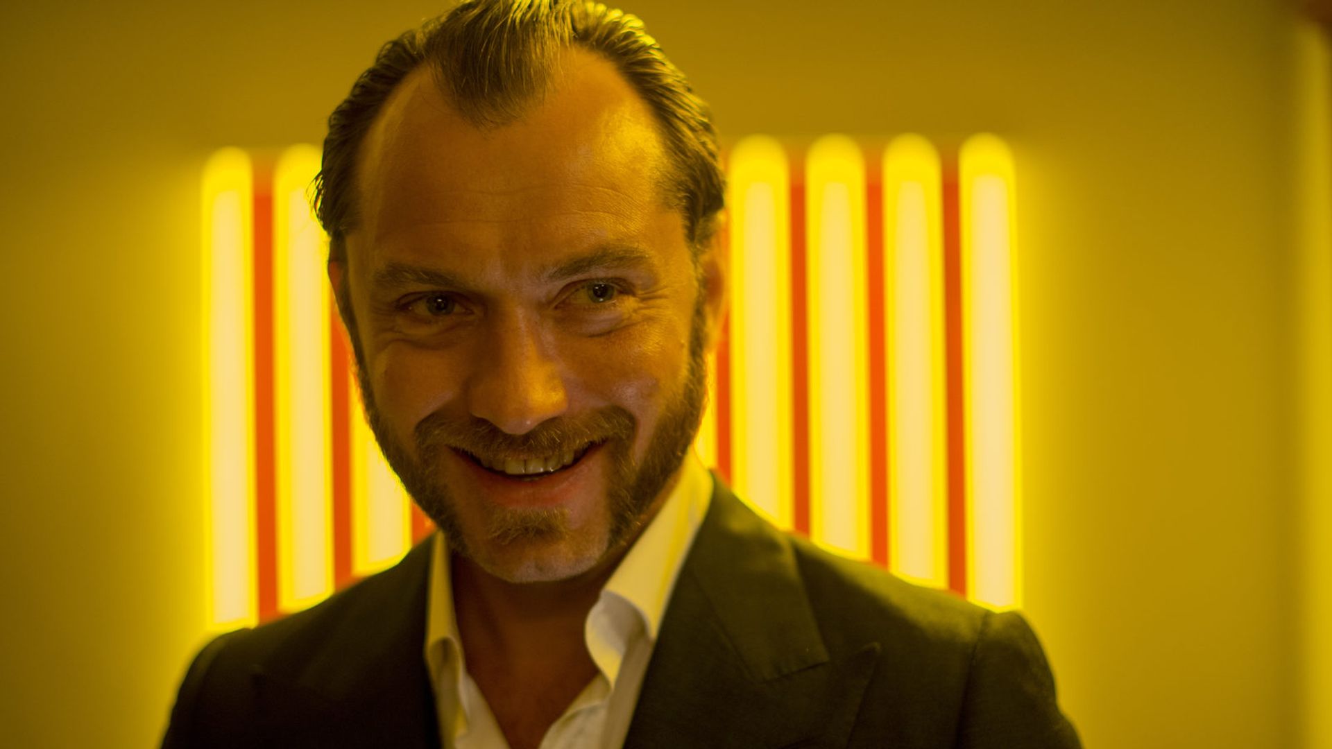 Jude Law con chaqueta sonríe contra un fondo de rayas de luz amarilla brillante