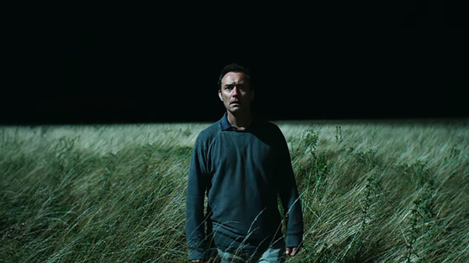 Jude Law con ropa oscura de pie en un campo de hierba alta por la noche, iluminado por una luz intensa