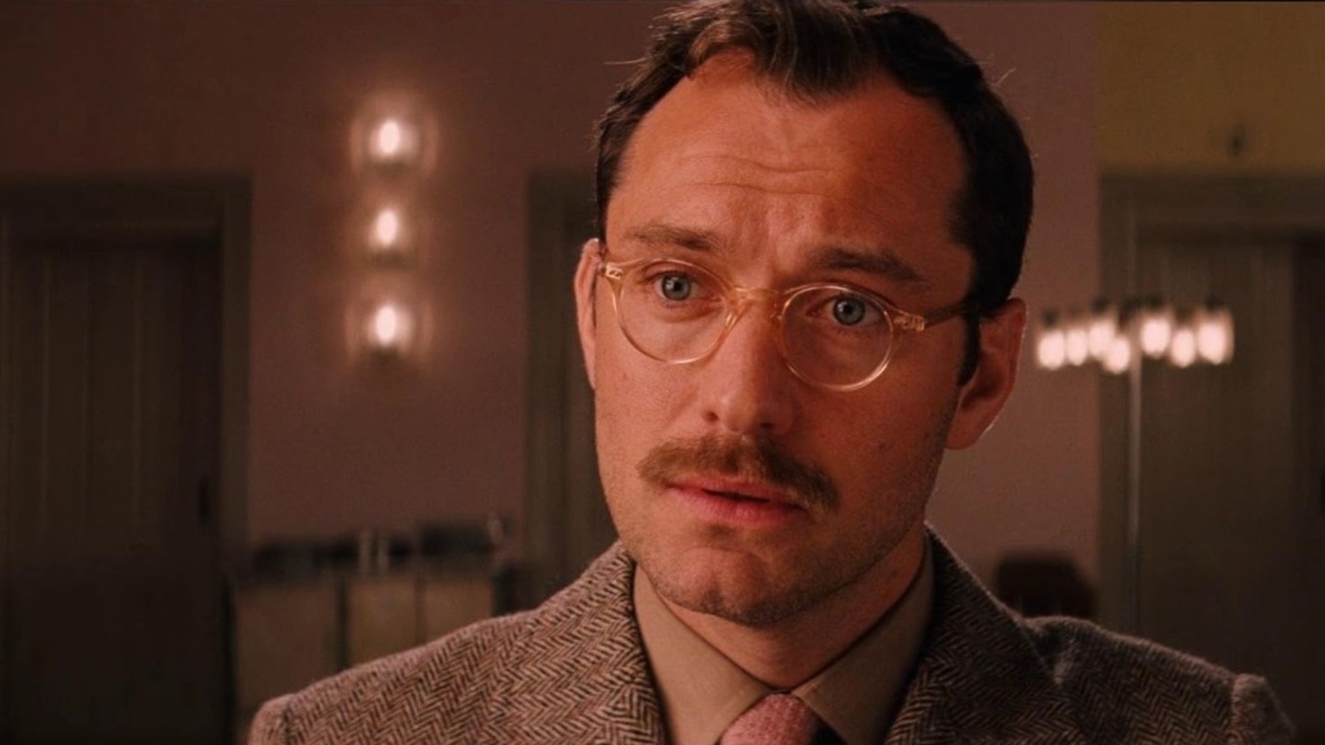 Jude Law con gafas y bigote en traje mira con expresión seria en el interior cálido de una habitación