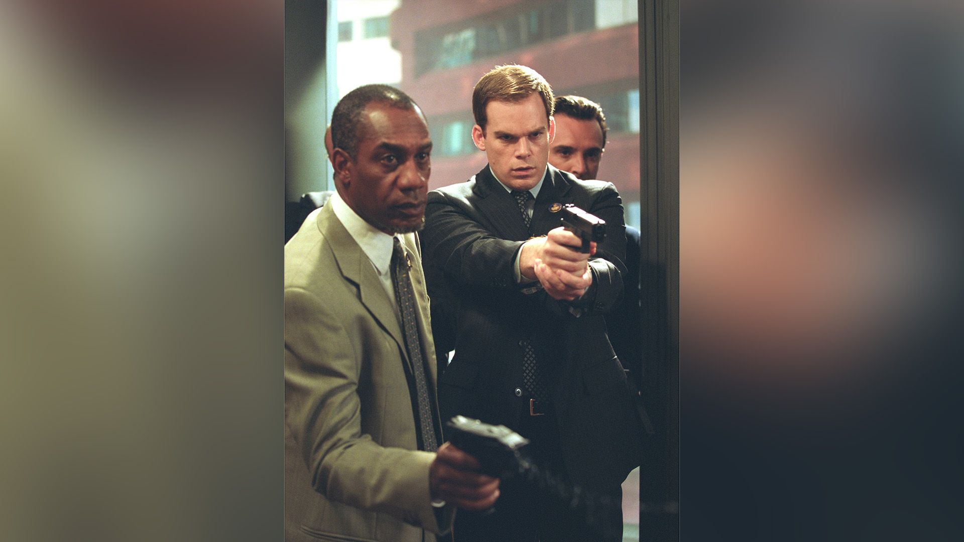 Michael C. Hall y un hombre negro de pie con los brazos extendidos sosteniendo armas
