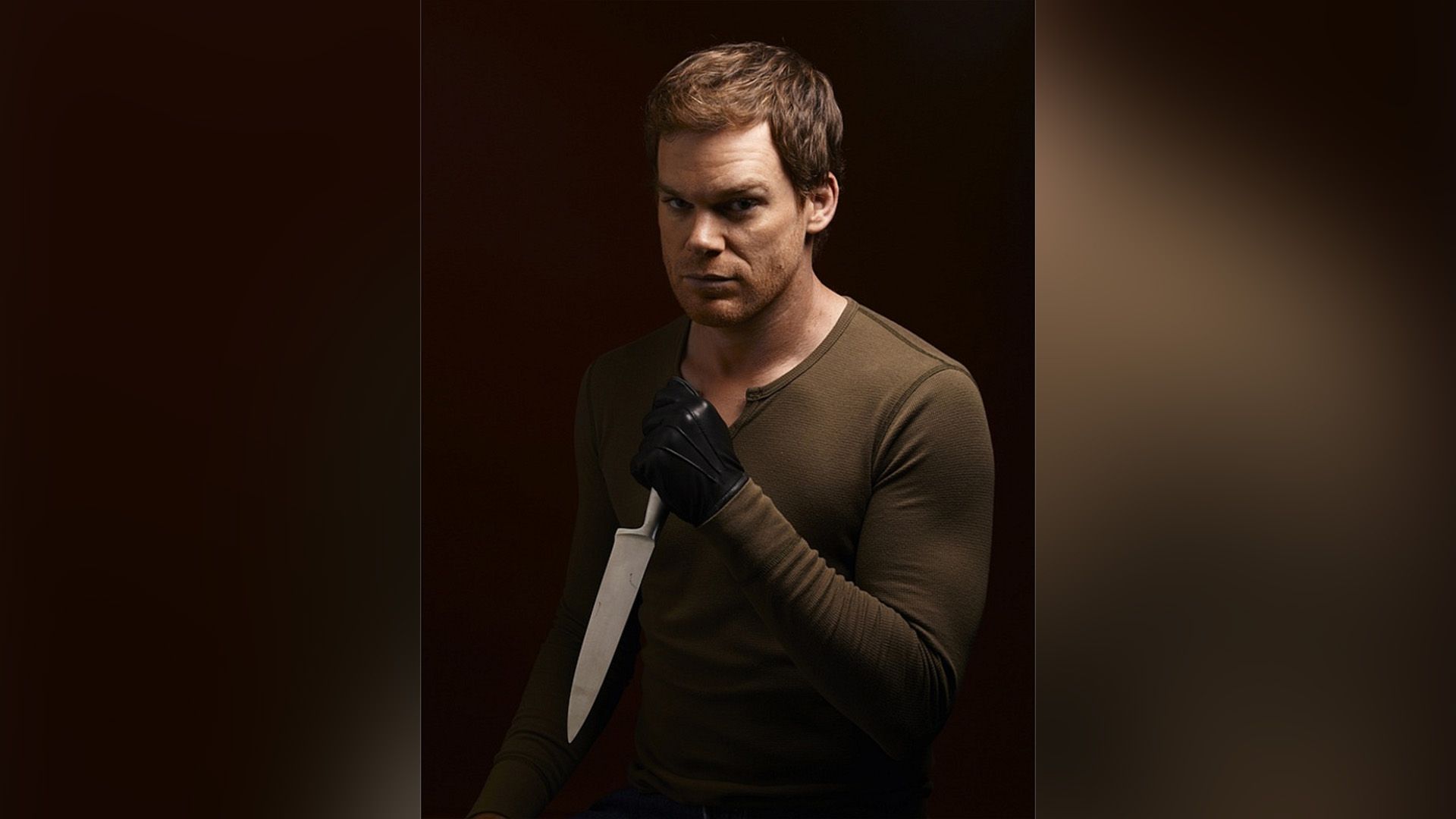 Michael C. Hall con una chaqueta marrón y guantes negros sosteniendo un cuchillo en la mano