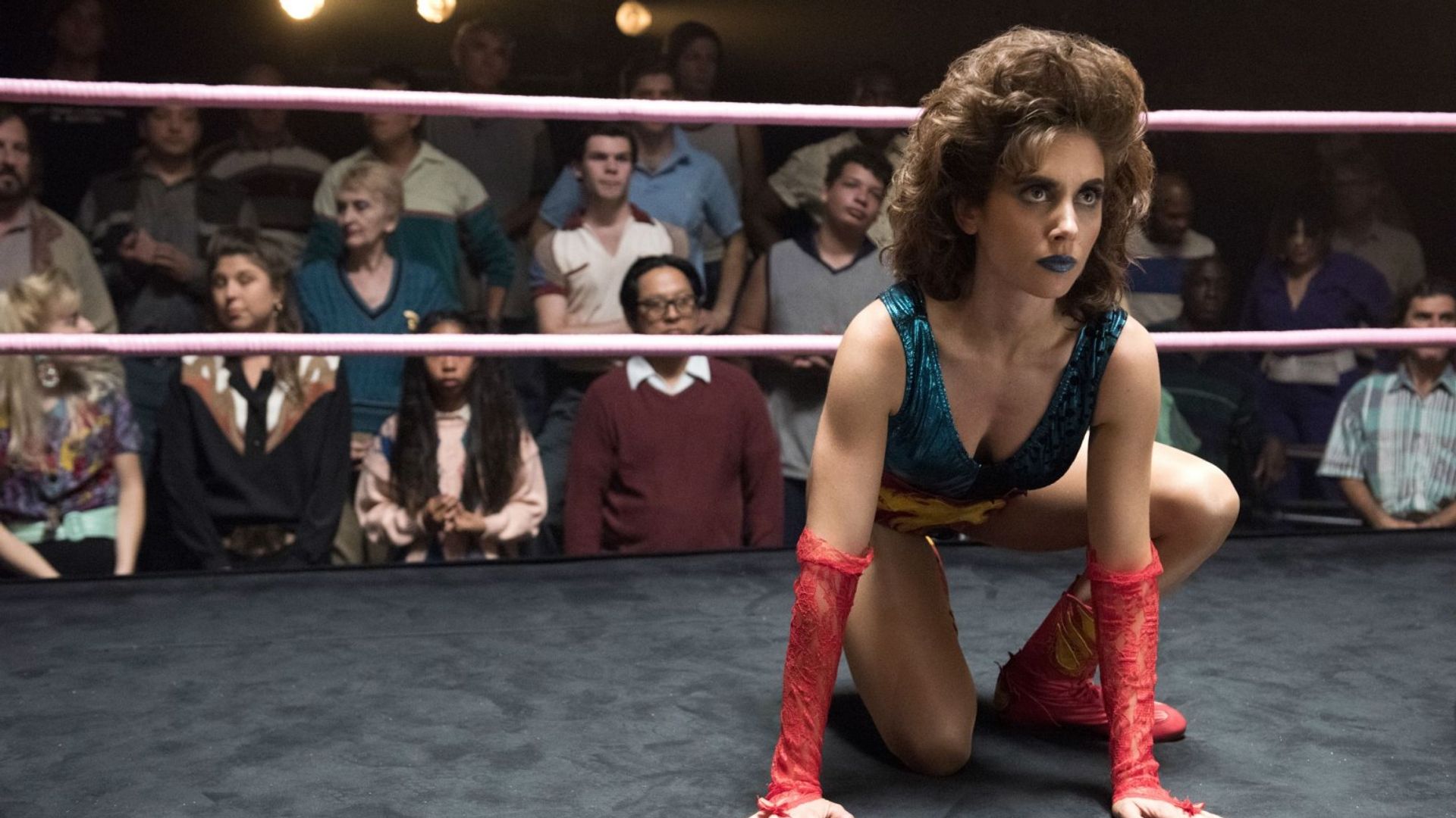 Alison Brie en traje de luchadora sentada en posición en el ring