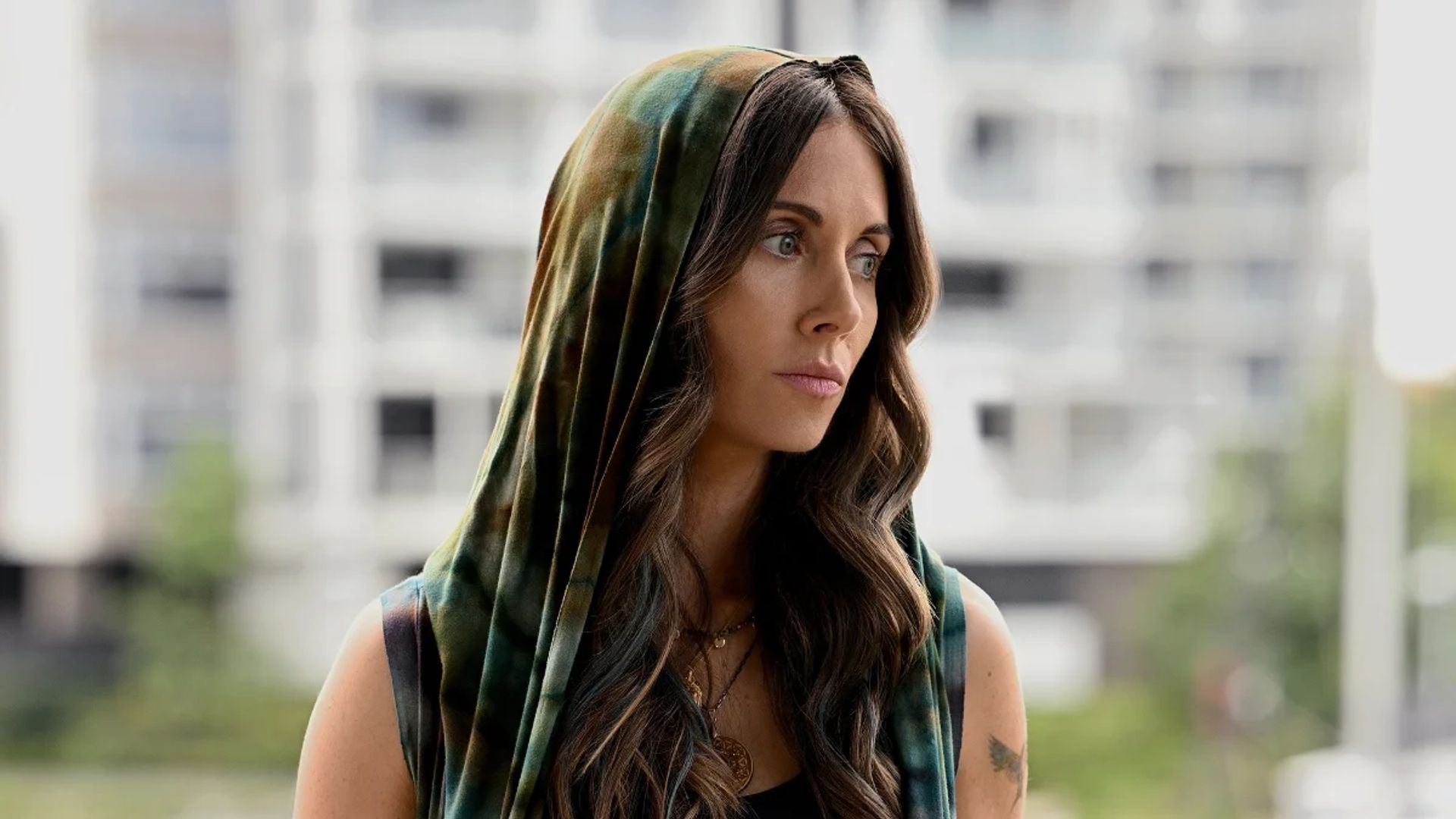 Alison Brie con cabello largo y una capucha mira pensativamente hacia un lado contra el telón de fondo de un edificio alto